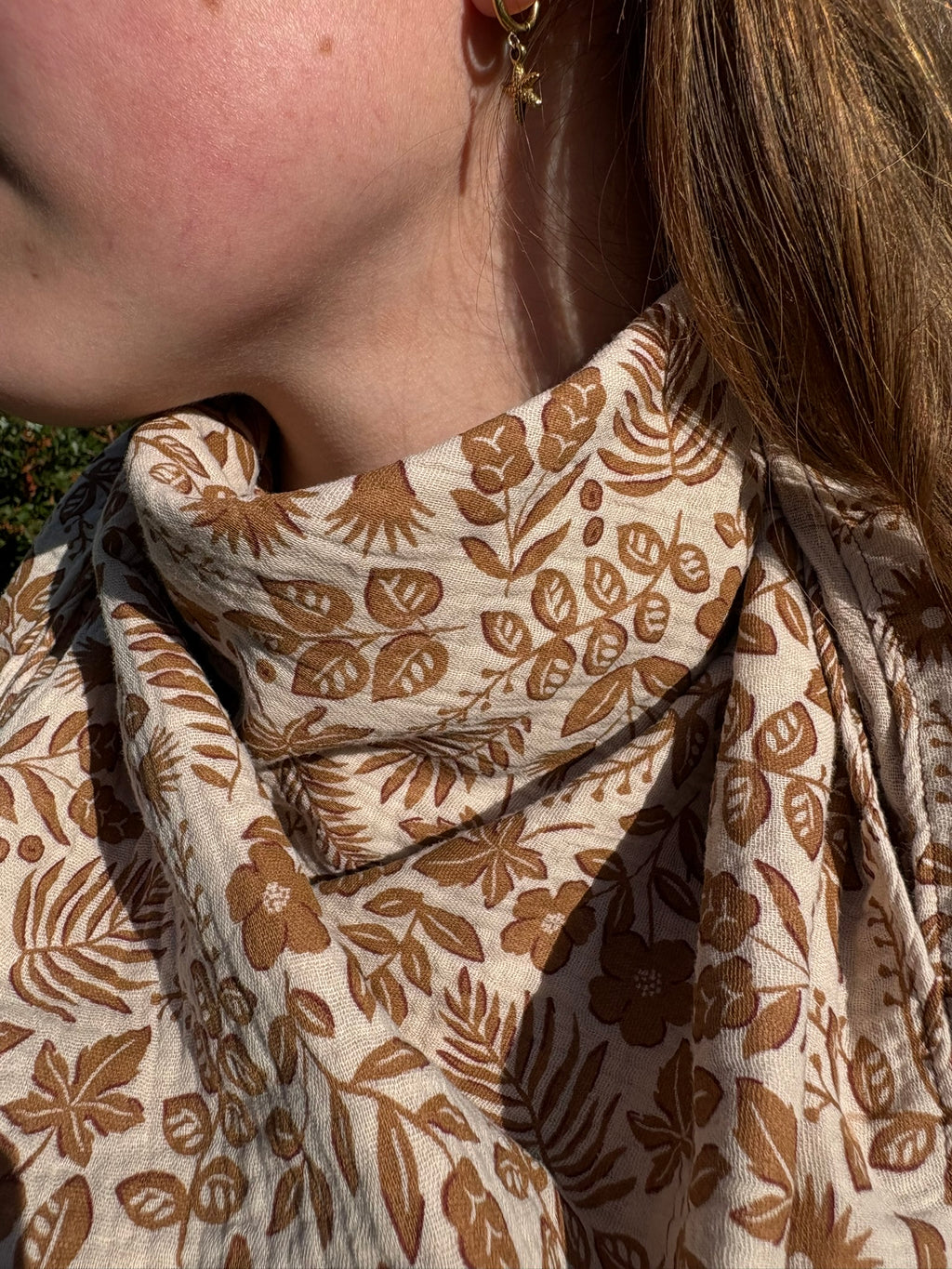 Chèche triangle en gaze de coton avec un motif végétal imprimé dans des tons de marron et beige, porté autour du cou par une jeune femme.