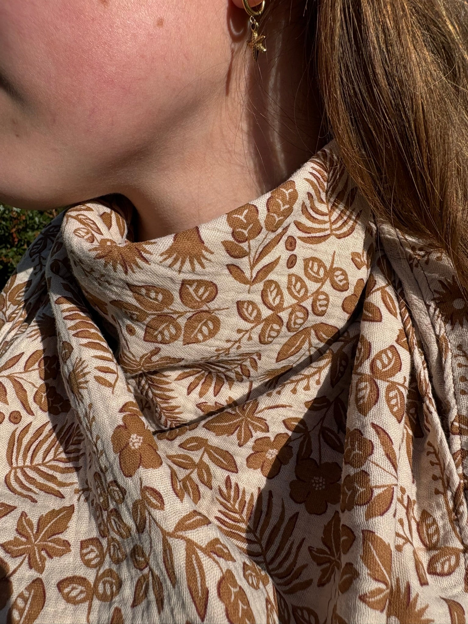 Chèche triangle en gaze de coton avec un motif végétal imprimé dans des tons de marron et beige, porté autour du cou par une jeune femme.