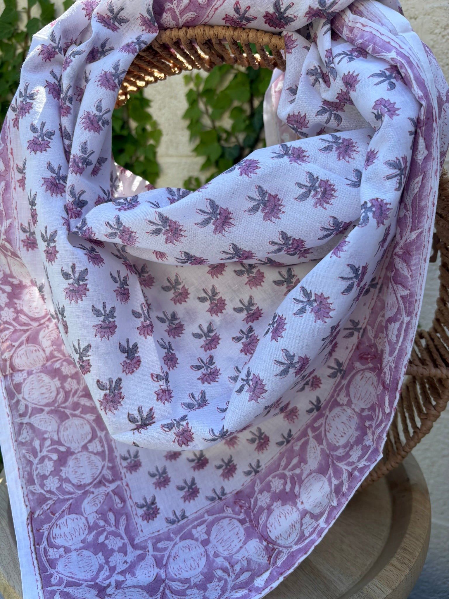 Un foulard en coton léger, orné de motifs artisanaux blockprint (impression au tampon de bois) dans des tons de vieux rose et parme sur fond blanc, est drapé artistiquement sur une chaise en osier tissé. Le foulard montre des détails subtils des motifs floraux et de la bordure travaillée. L'arrière-plan présente un mur ensoleillé et des plantes grimpantes vertes, suggérant une atmosphère naturelle et bohème.