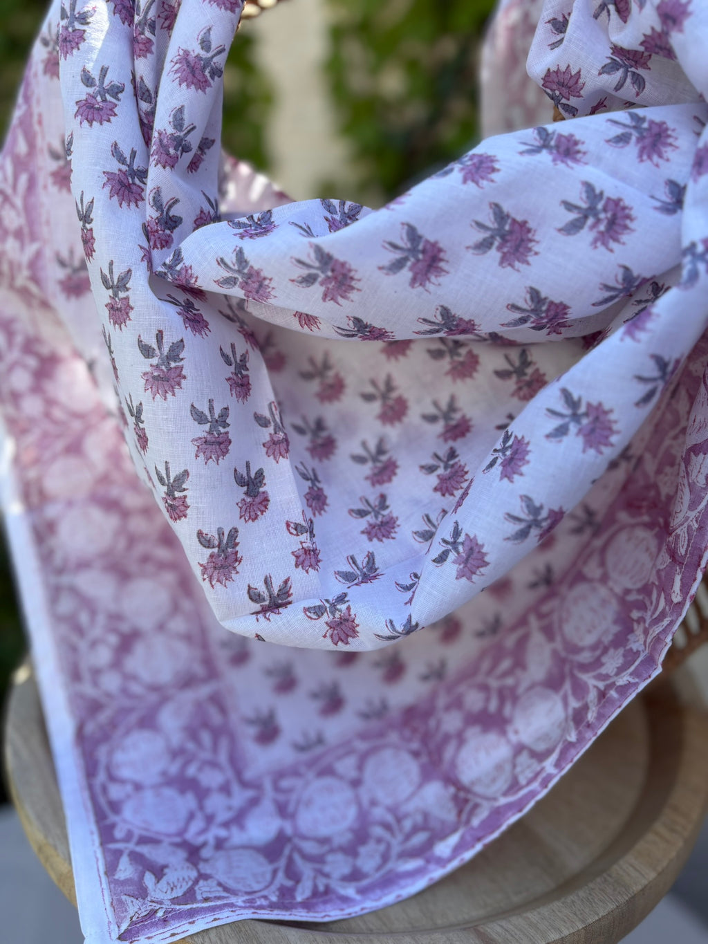 Un foulard en coton léger, orné de motifs artisanaux blockprint (impression au tampon de bois) dans des tons de vieux rose et parme sur fond blanc, est drapé artistiquement sur une chaise en osier tissé. Le foulard montre des détails subtils des motifs floraux et de la bordure travaillée. L'arrière-plan présente un mur ensoleillé et des plantes grimpantes vertes, suggérant une atmosphère naturelle et bohème.