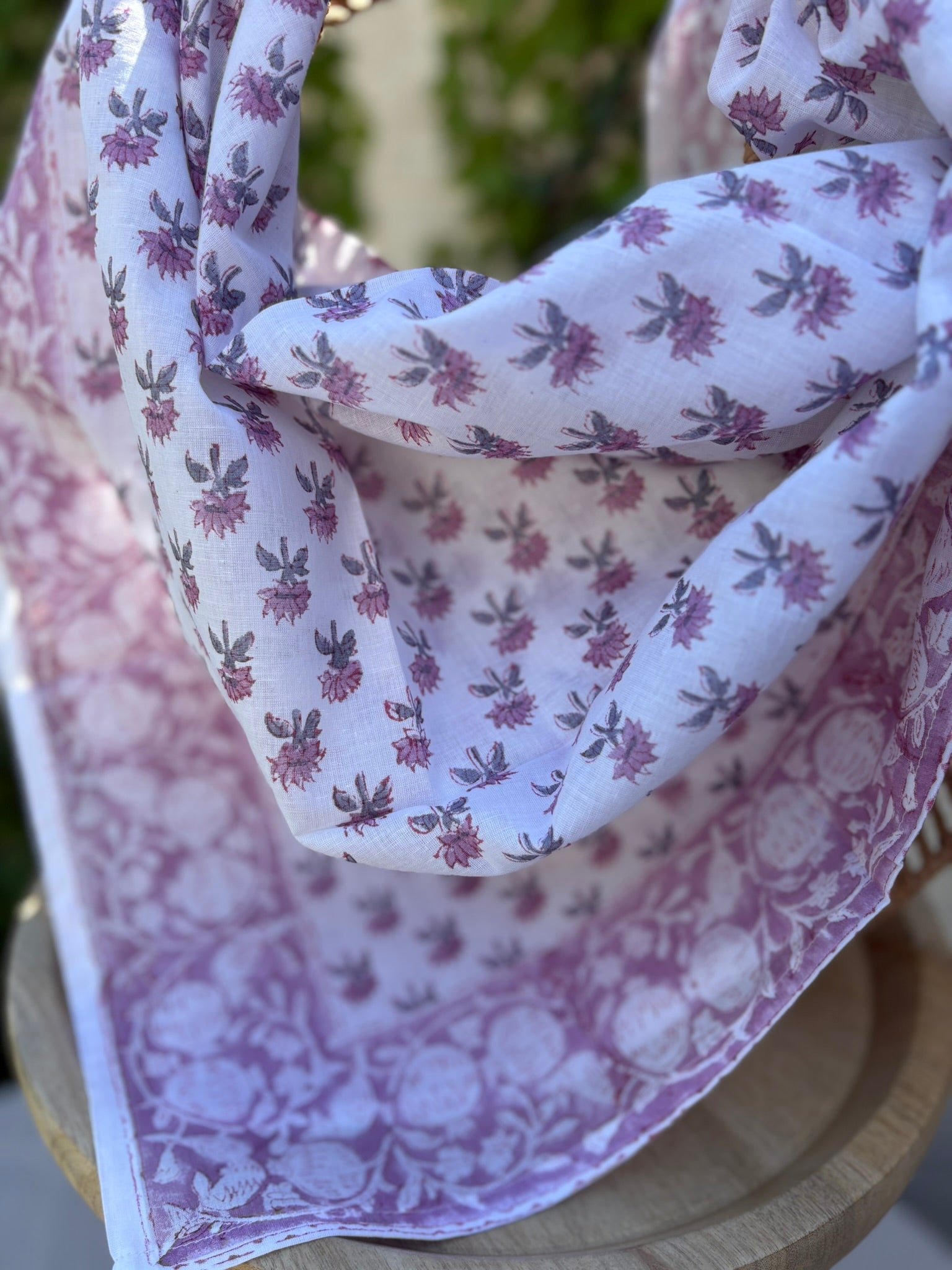 Un foulard en coton léger, orné de motifs artisanaux blockprint (impression au tampon de bois) dans des tons de vieux rose et parme sur fond blanc, est drapé artistiquement sur une chaise en osier tissé. Le foulard montre des détails subtils des motifs floraux et de la bordure travaillée. L'arrière-plan présente un mur ensoleillé et des plantes grimpantes vertes, suggérant une atmosphère naturelle et bohème.