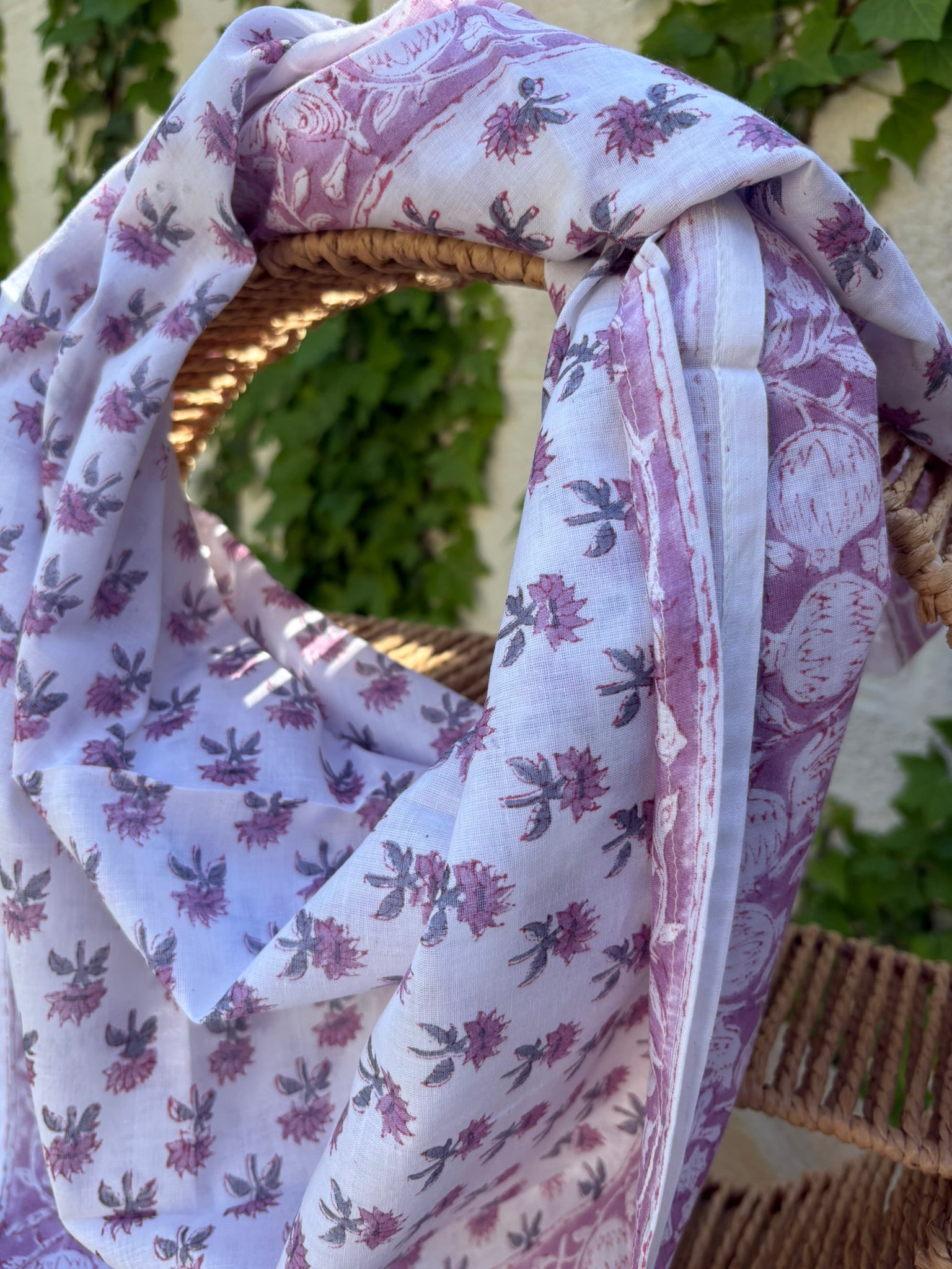 Un foulard en coton léger, orné de motifs artisanaux blockprint (impression au tampon de bois) dans des tons de vieux rose et parme sur fond blanc, est drapé artistiquement sur une chaise en osier tissé. Le foulard montre des détails subtils des motifs floraux et de la bordure travaillée. L'arrière-plan présente un mur ensoleillé et des plantes grimpantes vertes, suggérant une atmosphère naturelle et bohème.