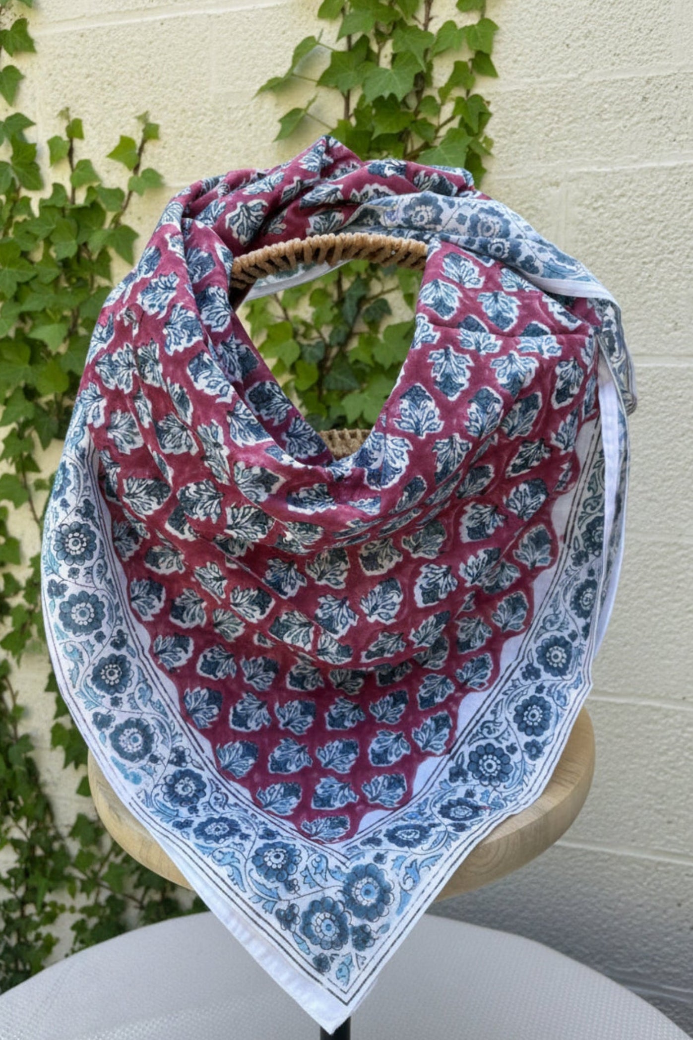 Un grand foulard en coton léger est drapé artistiquement sur un support en bois. Le motif central du foulard présente des feuillages stylisés bleu canard et gris répétés sur un fond lie de vin (vieux rose foncé). Le foulard est encadré par une large bordure blanche ornée de fleurs indiennes complexes et de motifs de médaillons, imprimés dans les mêmes tons de bleu et de lie de vin. La technique est de l'impression artisanale blockprint.  