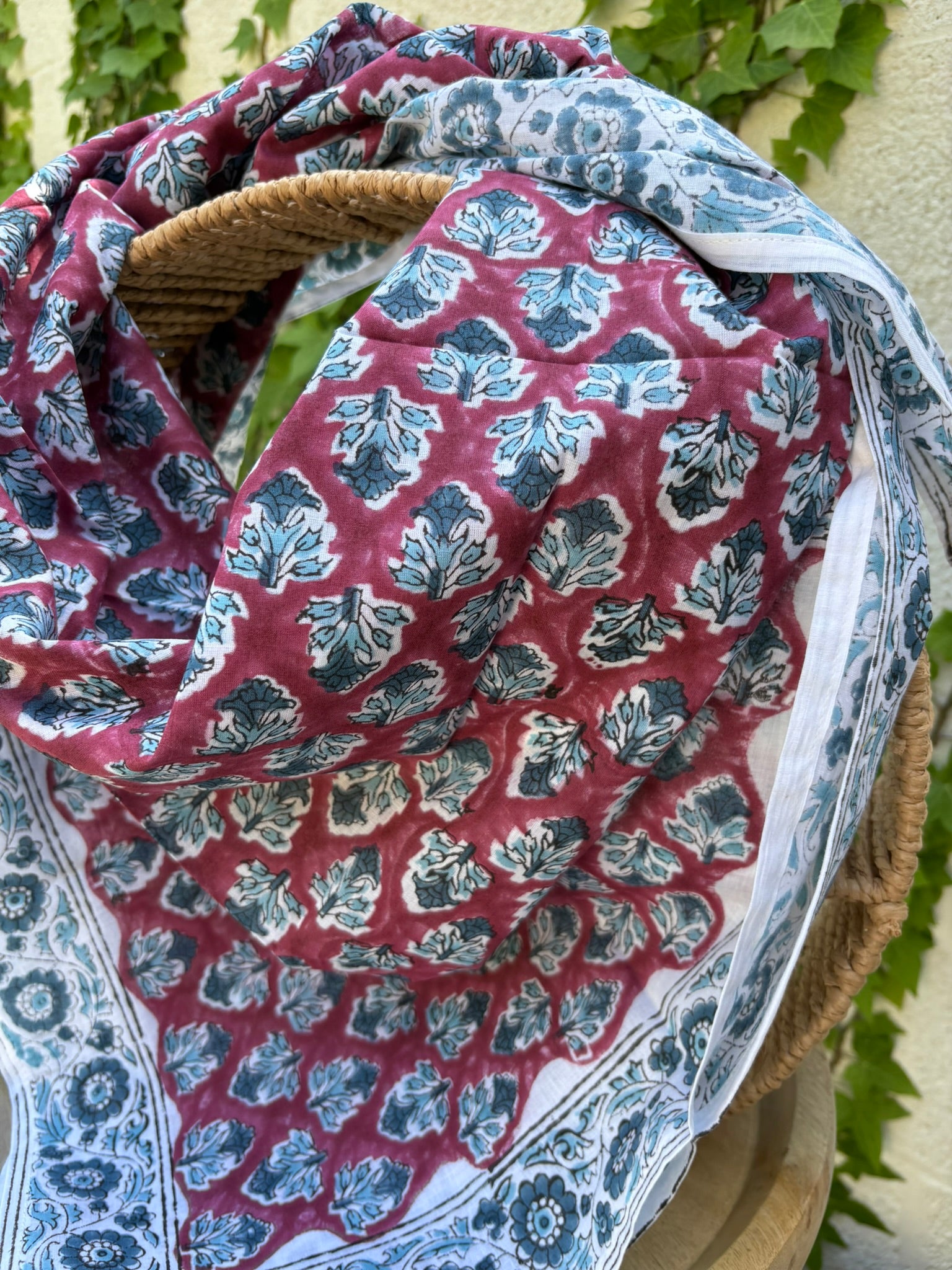 Un grand foulard en coton léger est drapé artistiquement sur un support en bois. Le motif central du foulard présente des feuillages stylisés bleu canard et gris répétés sur un fond lie de vin (vieux rose foncé). Le foulard est encadré par une large bordure blanche ornée de fleurs indiennes complexes et de motifs de médaillons, imprimés dans les mêmes tons de bleu et de lie de vin. La technique est de l'impression artisanale blockprint.  