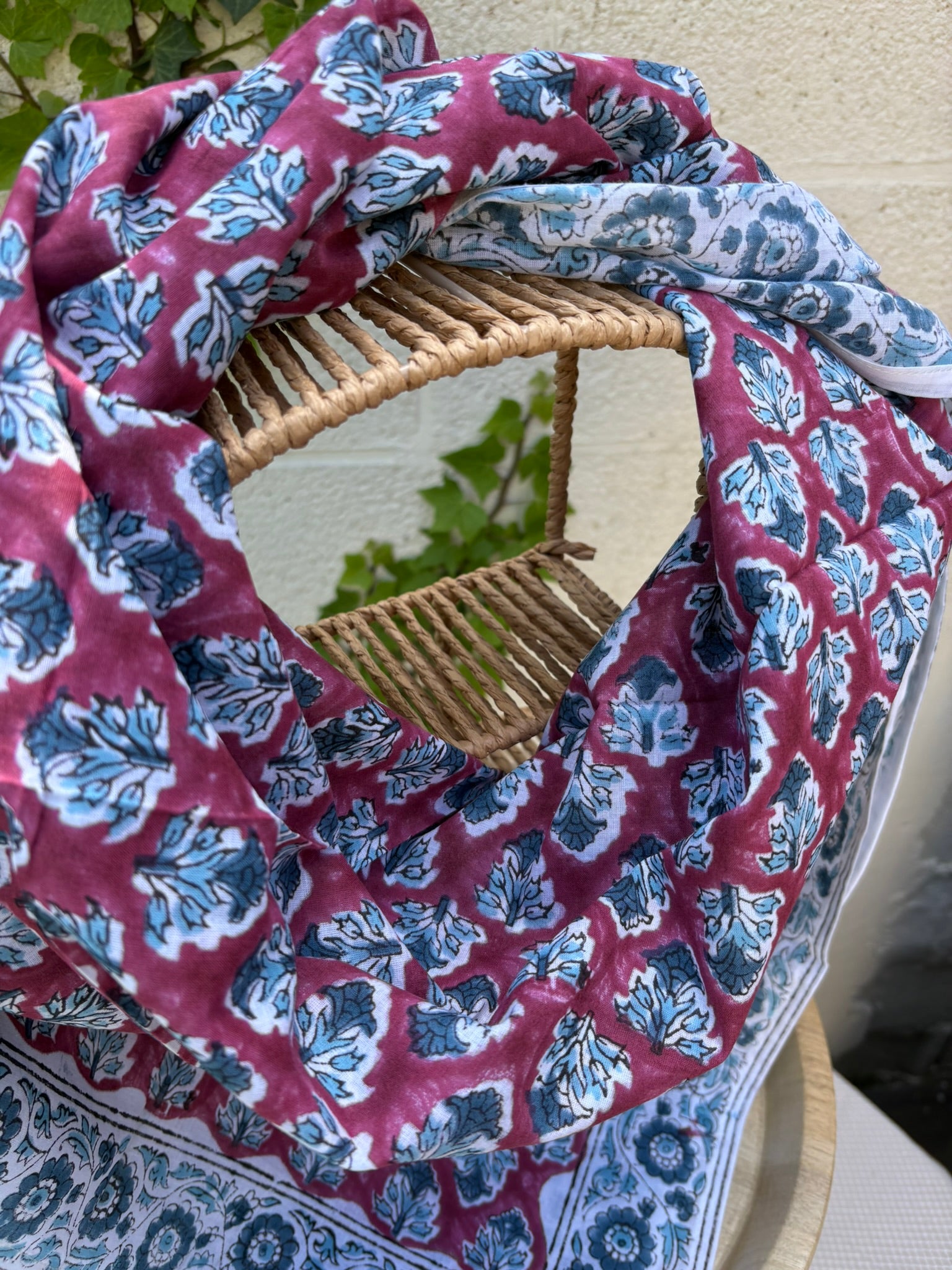 Un grand foulard en coton léger est drapé artistiquement sur un support en bois. Le motif central du foulard présente des feuillages stylisés bleu canard et gris répétés sur un fond lie de vin (vieux rose foncé). Le foulard est encadré par une large bordure blanche ornée de fleurs indiennes complexes et de motifs de médaillons, imprimés dans les mêmes tons de bleu et de lie de vin. La technique est de l'impression artisanale blockprint.  