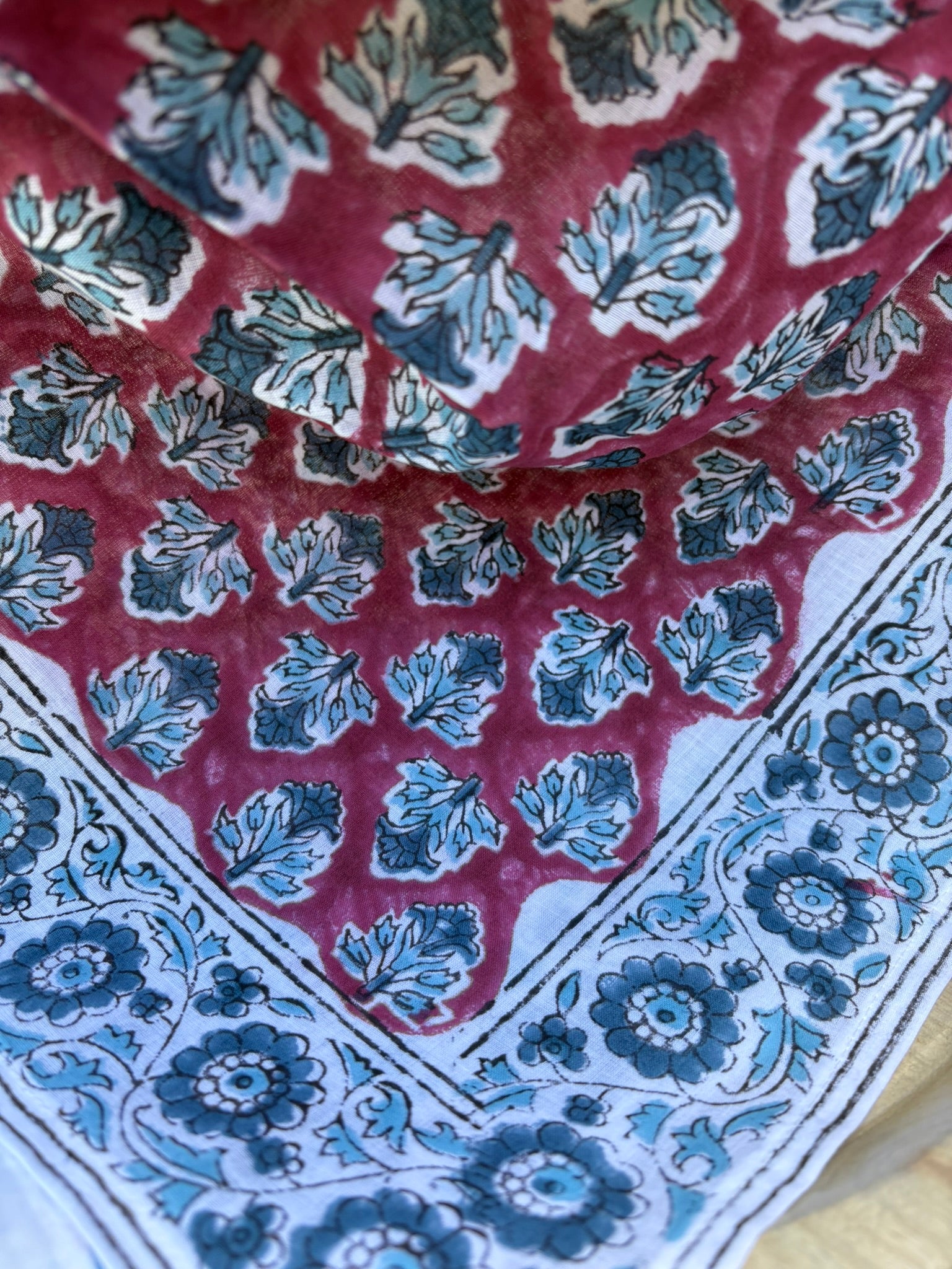 Un grand foulard en coton léger est drapé artistiquement sur un support en bois. Le motif central du foulard présente des feuillages stylisés bleu canard et gris répétés sur un fond lie de vin (vieux rose foncé). Le foulard est encadré par une large bordure blanche ornée de fleurs indiennes complexes et de motifs de médaillons, imprimés dans les mêmes tons de bleu et de lie de vin. La technique est de l'impression artisanale blockprint.  