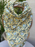 Un foulard artisanal en coton léger, présentant un imprimé blockprint floral, est mis en scène sur une chaise en osier circulaire. Le motif se compose de fleurs stylisées vert menthe et blanc, reliées par des tiges et des feuilles vert forêt foncé sur un fond jaune lumineux. Le foulard possède une bordure blanche délicate avec des motifs géométriques fins. L'image, prise en extérieur, montre un arrière-plan flou de lierre vert sur un mur clair, renforçant l'esthétique bohème et naturelle de l'accessoire