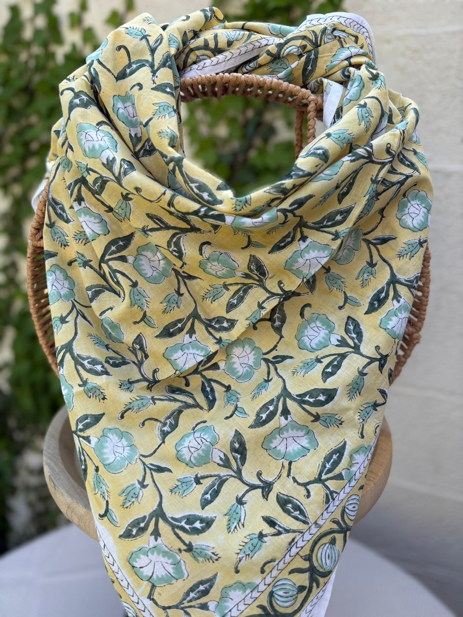 Un foulard artisanal en coton léger, présentant un imprimé blockprint floral, est mis en scène sur une chaise en osier circulaire. Le motif se compose de fleurs stylisées vert menthe et blanc, reliées par des tiges et des feuilles vert forêt foncé sur un fond jaune lumineux. Le foulard possède une bordure blanche délicate avec des motifs géométriques fins. L'image, prise en extérieur, montre un arrière-plan flou de lierre vert sur un mur clair, renforçant l'esthétique bohème et naturelle de l'accessoire