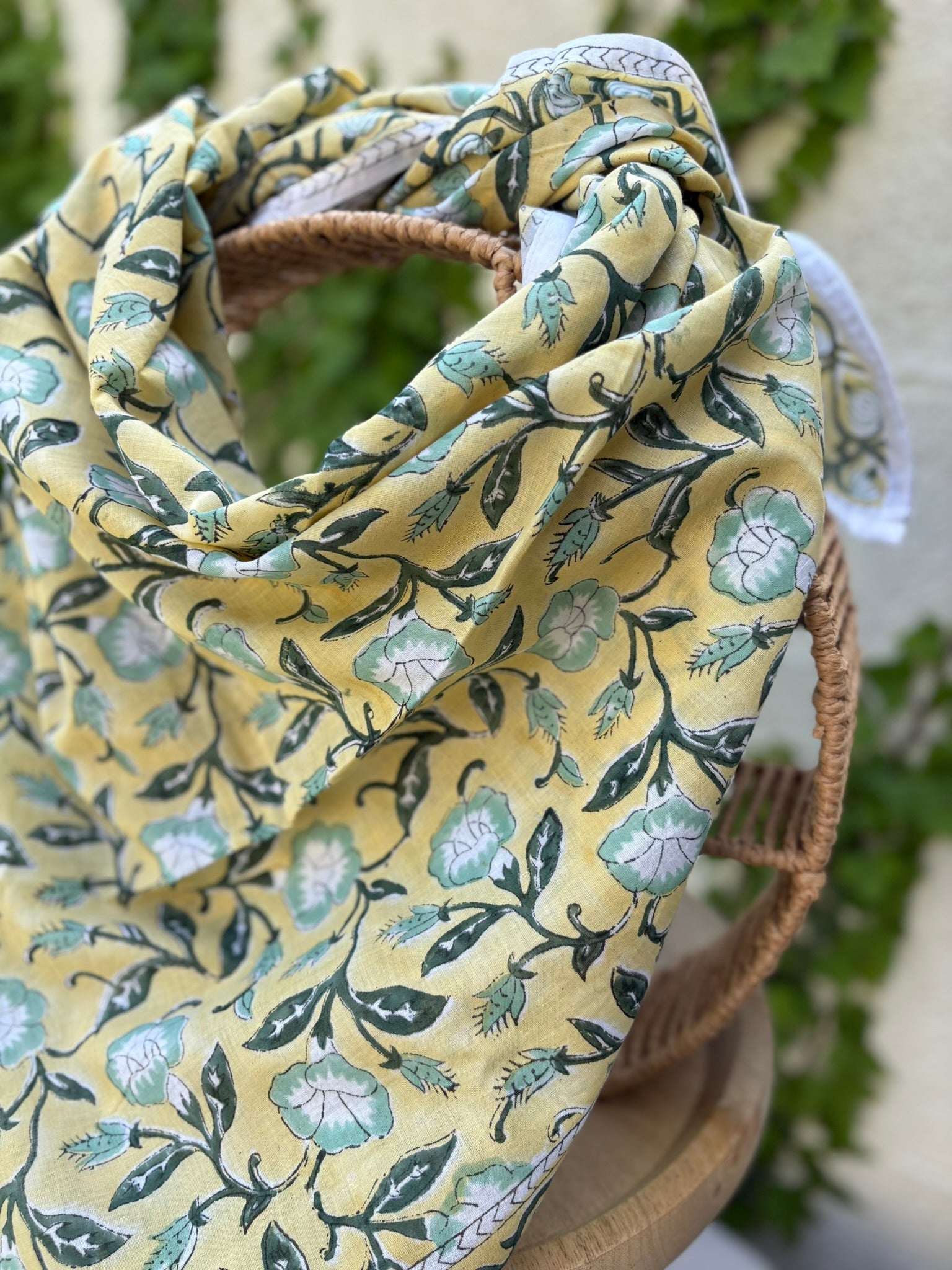 Un foulard artisanal en coton léger, présentant un imprimé blockprint floral, est mis en scène sur une chaise en osier circulaire. Le motif se compose de fleurs stylisées vert menthe et blanc, reliées par des tiges et des feuilles vert forêt foncé sur un fond jaune lumineux. Le foulard possède une bordure blanche délicate avec des motifs géométriques fins. L'image, prise en extérieur, montre un arrière-plan flou de lierre vert sur un mur clair, renforçant l'esthétique bohème et naturelle de l'accessoire
