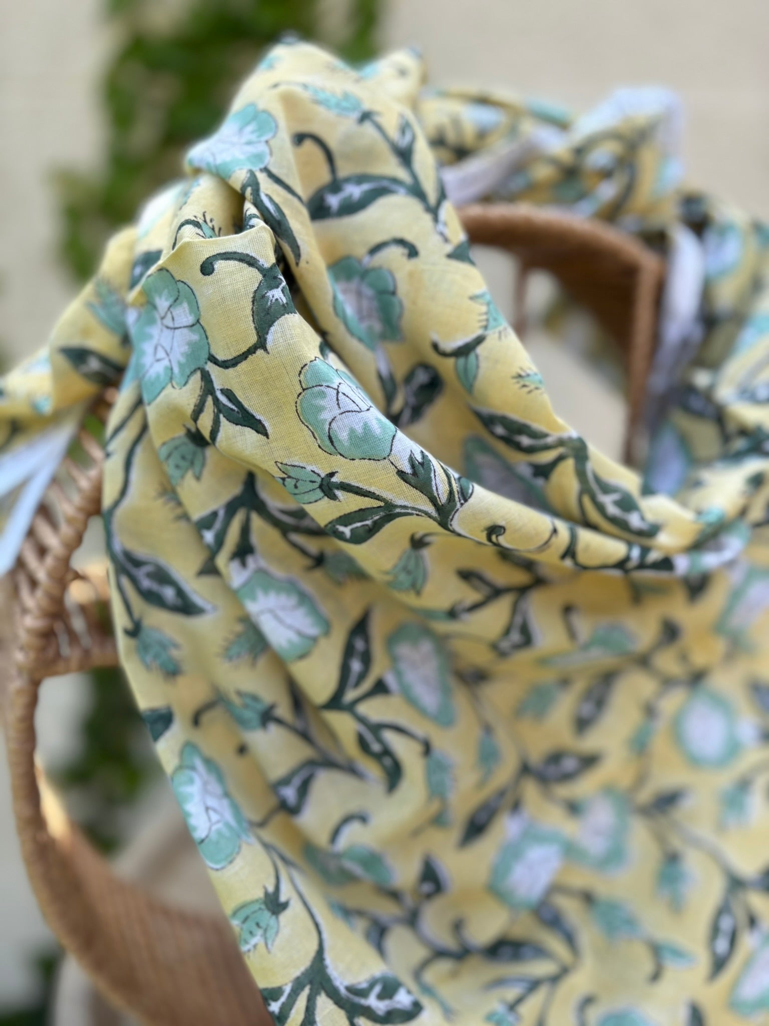 Un foulard artisanal en coton léger, présentant un imprimé blockprint floral, est mis en scène sur une chaise en osier circulaire. Le motif se compose de fleurs stylisées vert menthe et blanc, reliées par des tiges et des feuilles vert forêt foncé sur un fond jaune lumineux. Le foulard possède une bordure blanche délicate avec des motifs géométriques fins. L'image, prise en extérieur, montre un arrière-plan flou de lierre vert sur un mur clair, renforçant l'esthétique bohème et naturelle de l'accessoire