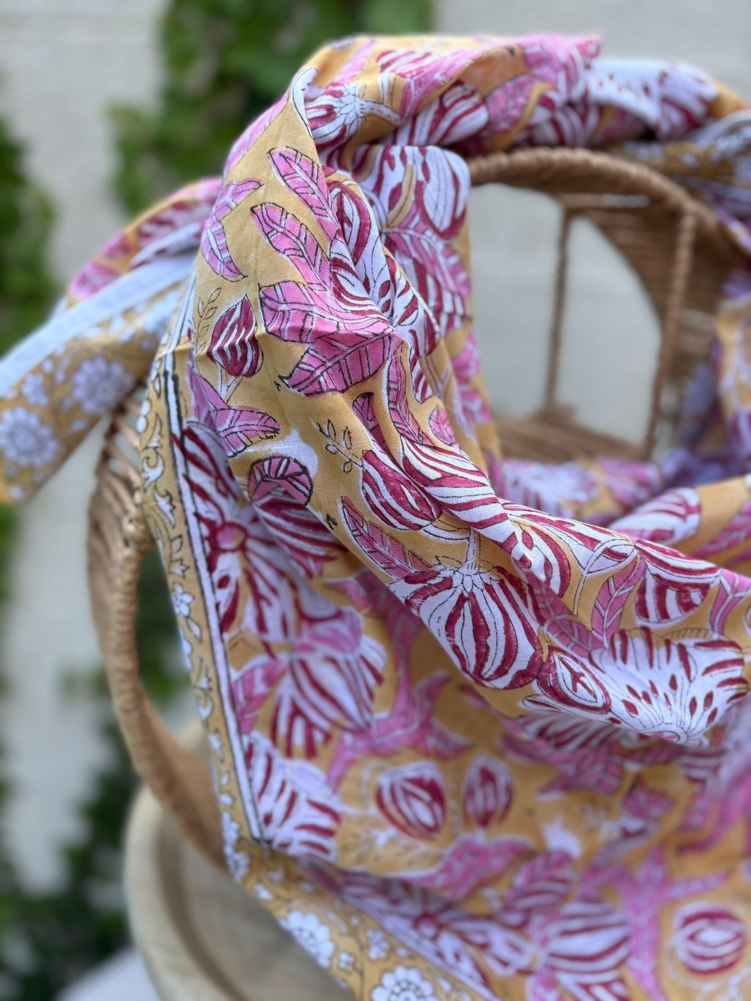 Un foulard en coton léger, imprimé selon la technique artisanale du blockprint, est présenté drapé sur une chaise en osier. Le design se compose de grandes fleurs épanouies et de feuilles striées dans des tons de rose framboise, bordeaux et blanc, le tout sur un fond jaune orangé chaleureux. Une bordure élégante sur fond blanc avec des motifs floraux noirs et jaunes vient terminer la pièce. L'arrière-plan, composé d'un mur clair et de lierre verdoyant, souligne l'aspect solaire et artisanal de l'accessoire.