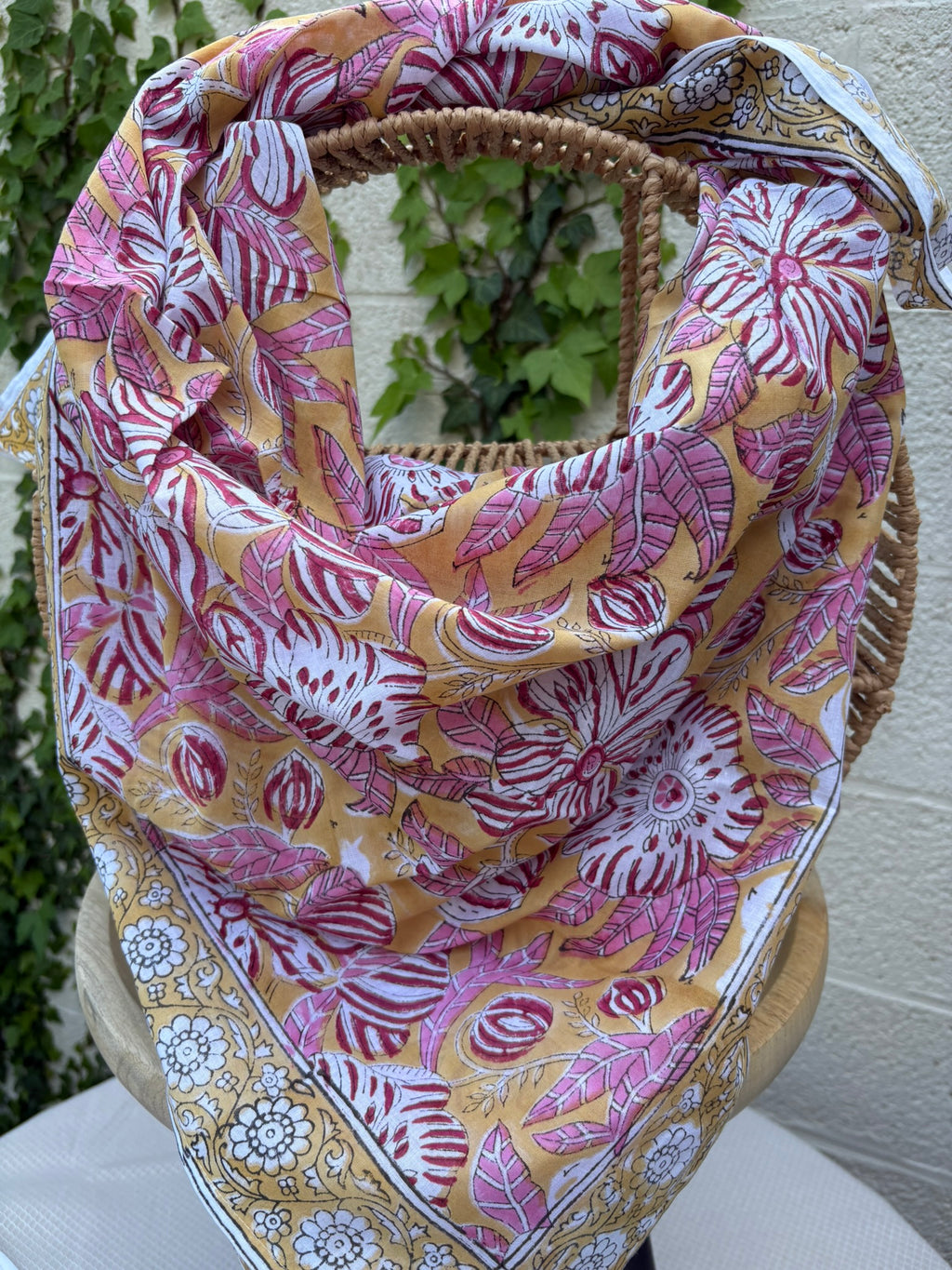 Un foulard en coton léger, imprimé selon la technique artisanale du blockprint, est présenté drapé sur une chaise en osier. Le design se compose de grandes fleurs épanouies et de feuilles striées dans des tons de rose framboise, bordeaux et blanc, le tout sur un fond jaune orangé chaleureux. Une bordure élégante sur fond blanc avec des motifs floraux noirs et jaunes vient terminer la pièce. L'arrière-plan, composé d'un mur clair et de lierre verdoyant, souligne l'aspect solaire et artisanal de l'accessoire.