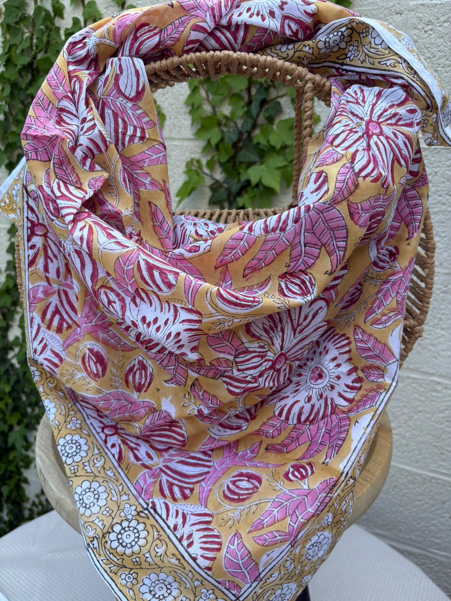 Un foulard en coton léger, imprimé selon la technique artisanale du blockprint, est présenté drapé sur une chaise en osier. Le design se compose de grandes fleurs épanouies et de feuilles striées dans des tons de rose framboise, bordeaux et blanc, le tout sur un fond jaune orangé chaleureux. Une bordure élégante sur fond blanc avec des motifs floraux noirs et jaunes vient terminer la pièce. L'arrière-plan, composé d'un mur clair et de lierre verdoyant, souligne l'aspect solaire et artisanal de l'accessoire.