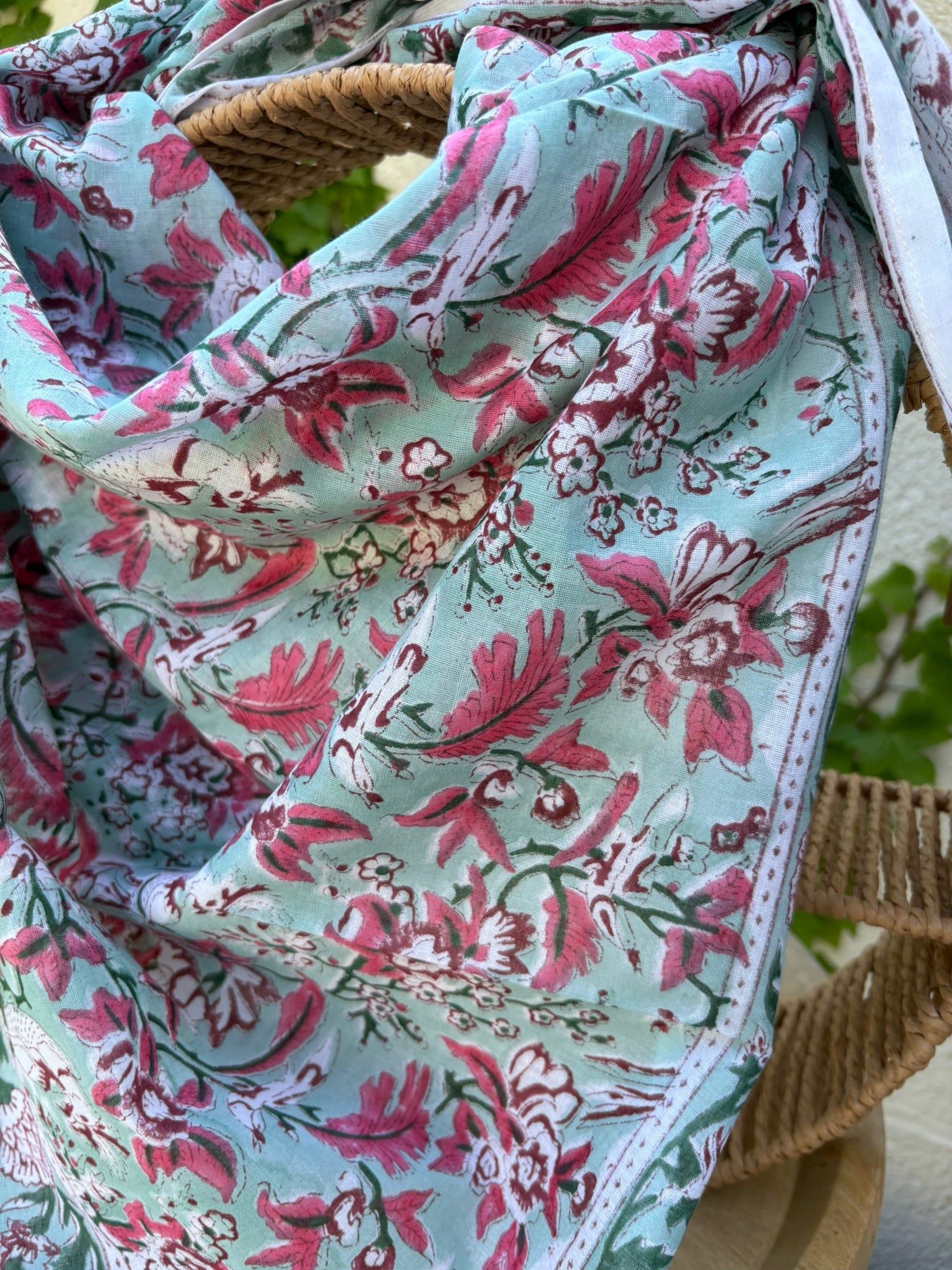 Un foulard en coton fin, réalisé avec la technique traditionnelle du blockprint, est présenté drapé sur une chaise en osier circulaire. Le motif se déploie sur un fond vert d'eau (céladon) et se compose de fleurs et de feuillages denses dans des tons de rose magenta, bordeaux et vert sapin. Une bordure blanche nette entoure le design floral. La mise en scène en extérieur, avec un mur clair et du lierre grimpant en arrière-plan, met en valeur les couleurs fraîches et le style bohème de l'accessoire.