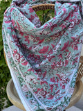 Un foulard en coton fin, réalisé avec la technique traditionnelle du blockprint, est présenté drapé sur une chaise en osier circulaire. Le motif se déploie sur un fond vert d'eau (céladon) et se compose de fleurs et de feuillages denses dans des tons de rose magenta, bordeaux et vert sapin. Une bordure blanche nette entoure le design floral. La mise en scène en extérieur, avec un mur clair et du lierre grimpant en arrière-plan, met en valeur les couleurs fraîches et le style bohème de l'accessoire.