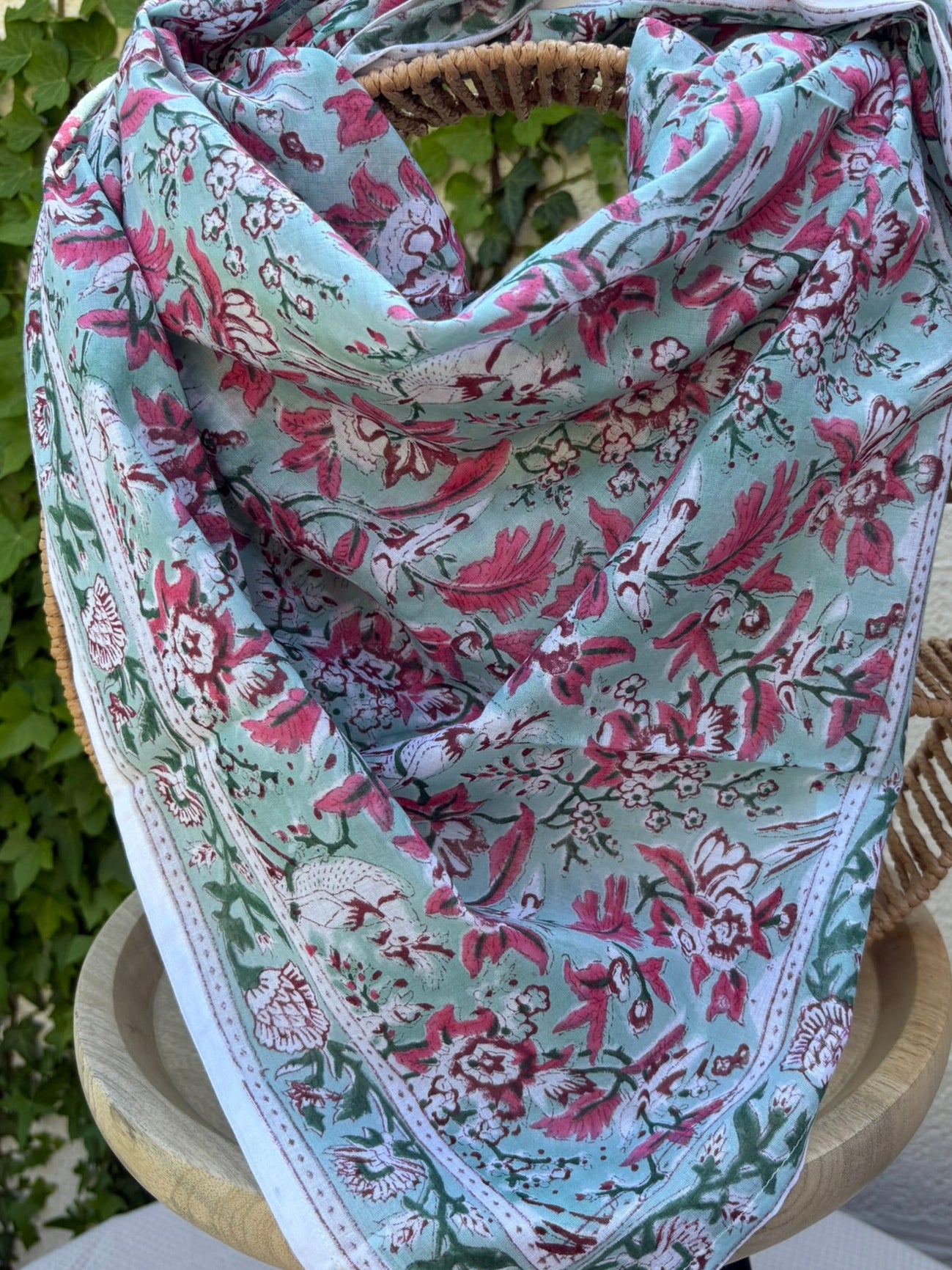 Un foulard en coton fin, réalisé avec la technique traditionnelle du blockprint, est présenté drapé sur une chaise en osier circulaire. Le motif se déploie sur un fond vert d'eau (céladon) et se compose de fleurs et de feuillages denses dans des tons de rose magenta, bordeaux et vert sapin. Une bordure blanche nette entoure le design floral. La mise en scène en extérieur, avec un mur clair et du lierre grimpant en arrière-plan, met en valeur les couleurs fraîches et le style bohème de l'accessoire.
