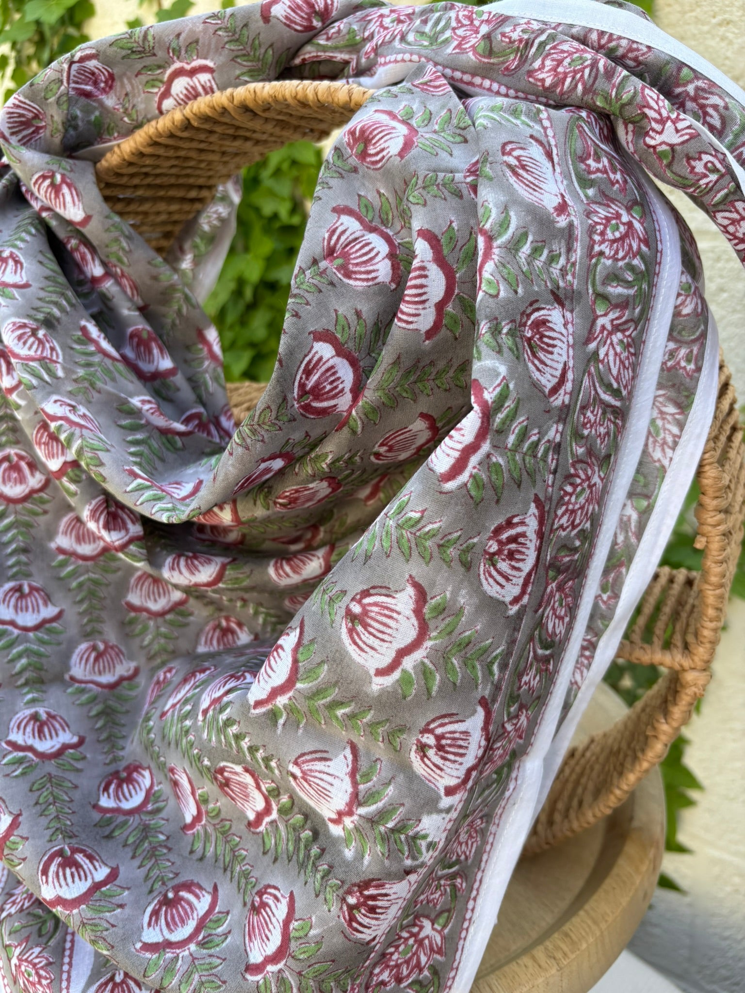 Un foulard en coton léger et fin, drapé artistiquement sur une chaise en osier. Le foulard, de couleur gris souris neutre, est entièrement recouvert d'un motif répétitif imprimé à la main (block print). Ce motif se compose de fleurs stylisées, rappelant des tulipes ou des bourgeons, dans des tons de rouge bordeaux et lie de vin, reliées par des tiges et des feuilles vert tendre disposées en chevrons. La technique d'impression artisanale laisse apparaître de légères irrégularités authentiques dans le dessin
