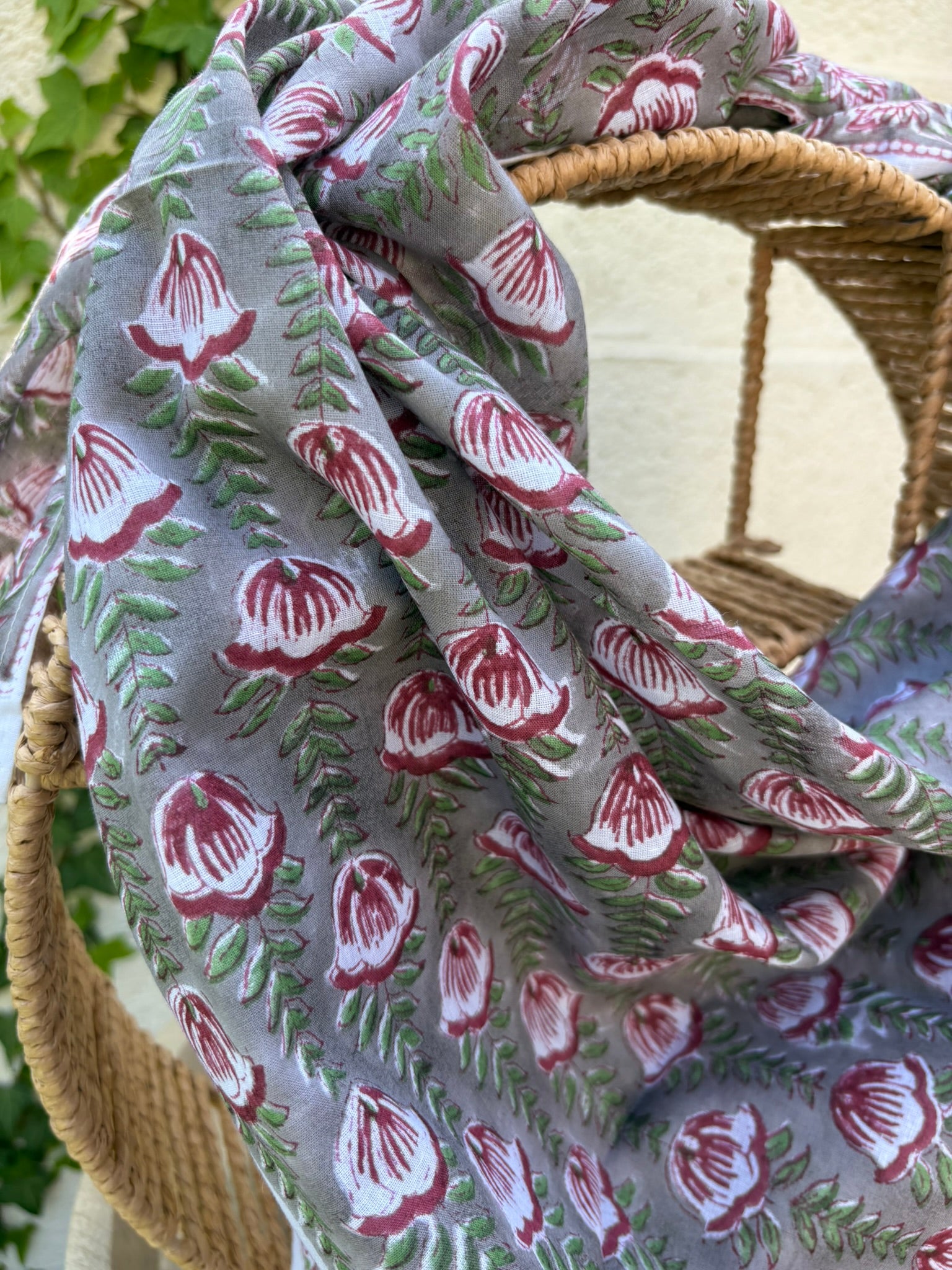 Un foulard en coton léger et fin, drapé artistiquement sur une chaise en osier. Le foulard, de couleur gris souris neutre, est entièrement recouvert d'un motif répétitif imprimé à la main (block print). Ce motif se compose de fleurs stylisées, rappelant des tulipes ou des bourgeons, dans des tons de rouge bordeaux et lie de vin, reliées par des tiges et des feuilles vert tendre disposées en chevrons. La technique d'impression artisanale laisse apparaître de légères irrégularités authentiques dans le dessin