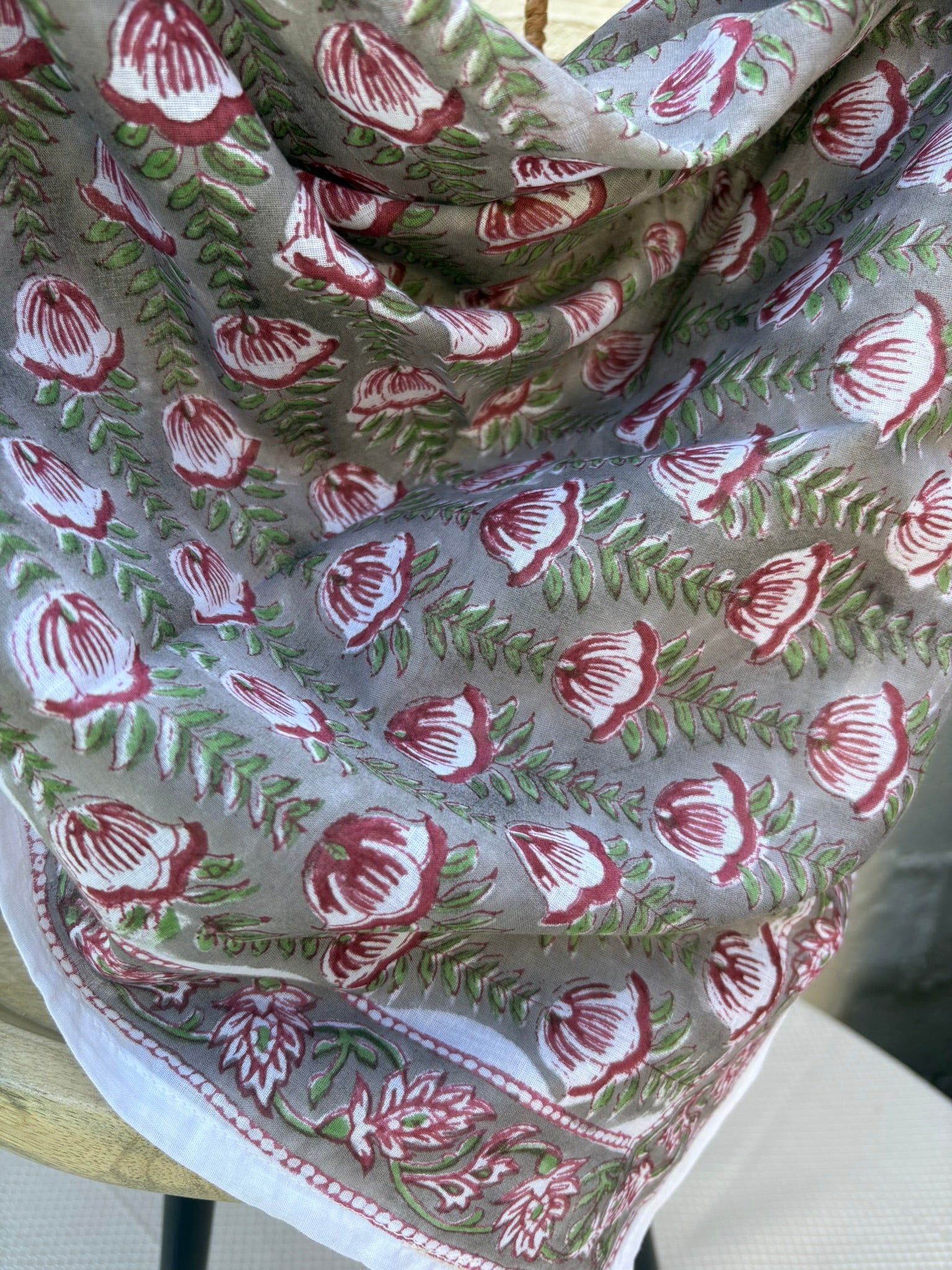 Un foulard en coton léger et fin, drapé artistiquement sur une chaise en osier. Le foulard, de couleur gris souris neutre, est entièrement recouvert d'un motif répétitif imprimé à la main (block print). Ce motif se compose de fleurs stylisées, rappelant des tulipes ou des bourgeons, dans des tons de rouge bordeaux et lie de vin, reliées par des tiges et des feuilles vert tendre disposées en chevrons. La technique d'impression artisanale laisse apparaître de légères irrégularités authentiques dans le dessin