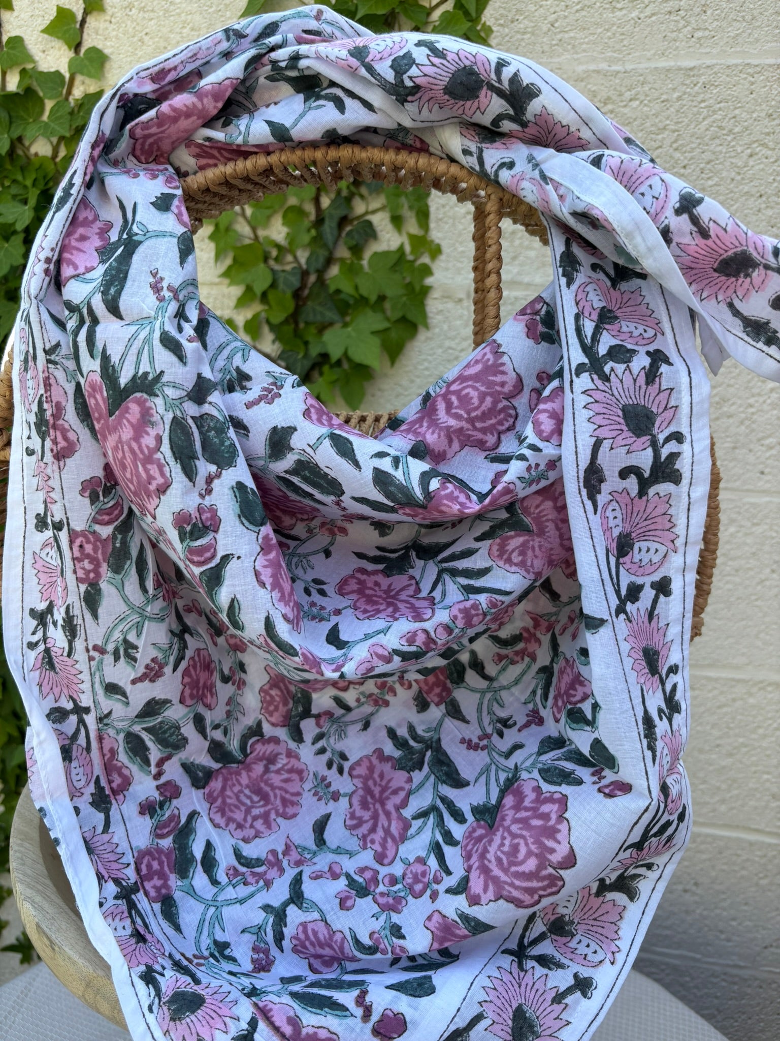 Un foulard en coton léger et fin est mis en scène, drapé artistiquement sur une chaise en osier tissé. Le foulard, de couleur blanc est orné d'un motif répétitif imprimé à la main (block print). Ce motif se compose de grandes fleurs épanouies, de feuillages et d'ornements stylisés dans des tons de vieux rose et de gris/vert. La technique d'impression artisanale laisse apparaître de légères irrégularités authentiques dans le dessin. Le foulard est plié et drapé de manière à montrer les plis et la fluidité