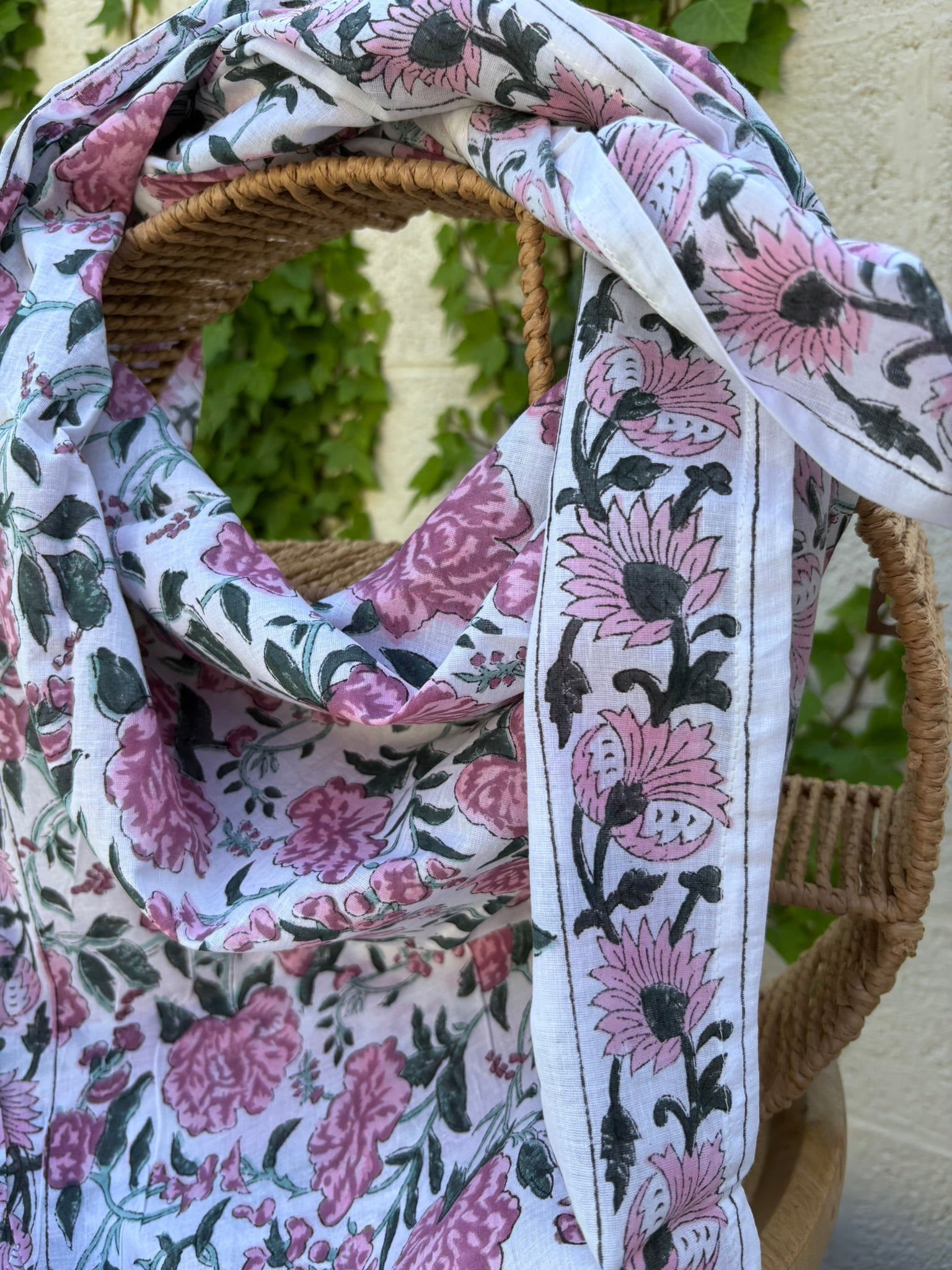 Un foulard en coton léger et fin est mis en scène, drapé artistiquement sur une chaise en osier tissé. Le foulard, de couleur blanc est orné d'un motif répétitif imprimé à la main (block print). Ce motif se compose de grandes fleurs épanouies, de feuillages et d'ornements stylisés dans des tons de vieux rose et de gris/vert. La technique d'impression artisanale laisse apparaître de légères irrégularités authentiques dans le dessin. Le foulard est plié et drapé de manière à montrer les plis et la fluidité