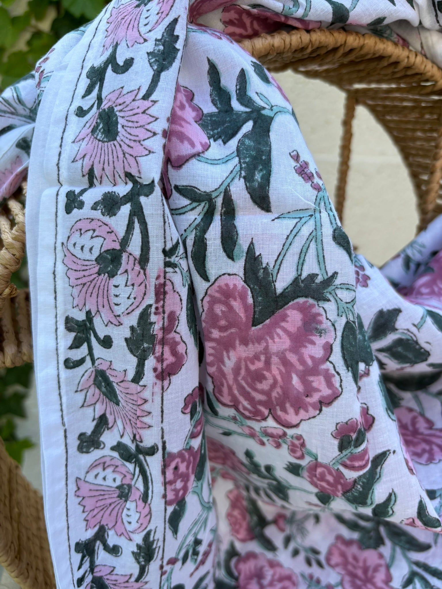 Un foulard en coton léger et fin est mis en scène, drapé artistiquement sur une chaise en osier tissé. Le foulard, de couleur blanc est orné d'un motif répétitif imprimé à la main (block print). Ce motif se compose de grandes fleurs épanouies, de feuillages et d'ornements stylisés dans des tons de vieux rose et de gris/vert. La technique d'impression artisanale laisse apparaître de légères irrégularités authentiques dans le dessin. Le foulard est plié et drapé de manière à montrer les plis et la fluidité