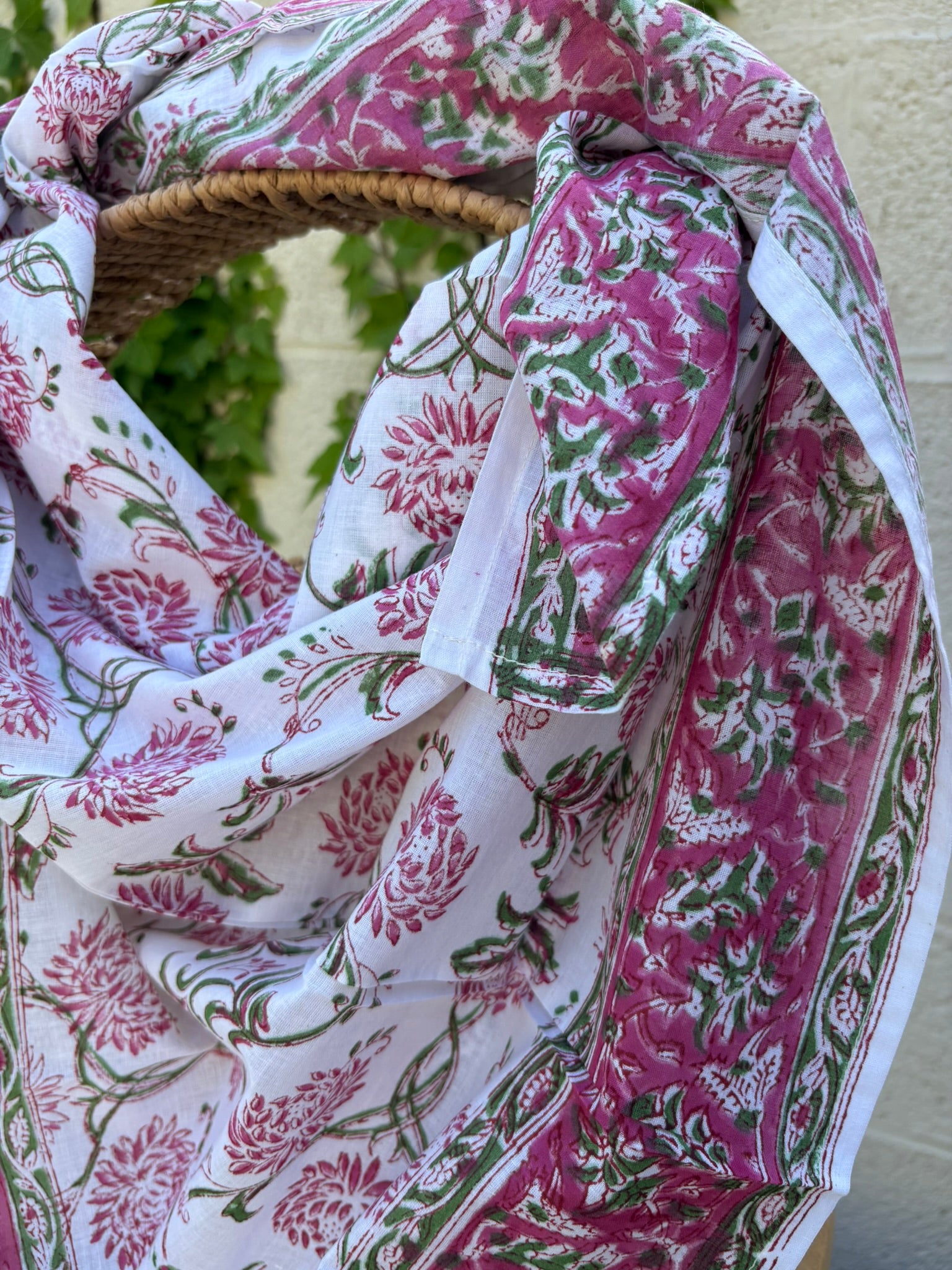Un foulard en voile de coton blanc, léger et aérien, est présenté drapé sur une chaise en osier. Il est orné d'un imprimé artisanal blockprint représentant des fleurs de lotus et de chrysanthèmes stylisées dans des tons rose fuchsia et rose indien, reliées par des tiges et des feuilles vert tendre. Une large bordure fleurie assortie encadre la pièce. La mise en scène, avec un arrière-plan flou composé de lierre vert et d'un mur clair, souligne le caractère naturel, lumineux et bohème de cet accessoire