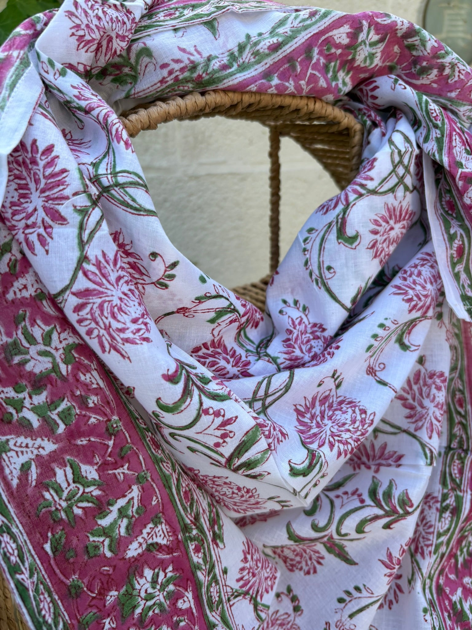 Un foulard en voile de coton blanc, léger et aérien, est présenté drapé sur une chaise en osier. Il est orné d'un imprimé artisanal blockprint représentant des fleurs de lotus et de chrysanthèmes stylisées dans des tons rose fuchsia et rose indien, reliées par des tiges et des feuilles vert tendre. Une large bordure fleurie assortie encadre la pièce. La mise en scène, avec un arrière-plan flou composé de lierre vert et d'un mur clair, souligne le caractère naturel, lumineux et bohème de cet accessoire