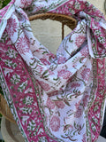 Un foulard en voile de coton blanc, léger et aérien, est présenté drapé sur une chaise en osier. Il est orné d'un imprimé artisanal blockprint représentant des fleurs de lotus et de chrysanthèmes stylisées dans des tons rose fuchsia et rose indien, reliées par des tiges et des feuilles vert tendre. Une large bordure fleurie assortie encadre la pièce. La mise en scène, avec un arrière-plan flou composé de lierre vert et d'un mur clair, souligne le caractère naturel, lumineux et bohème de cet accessoire