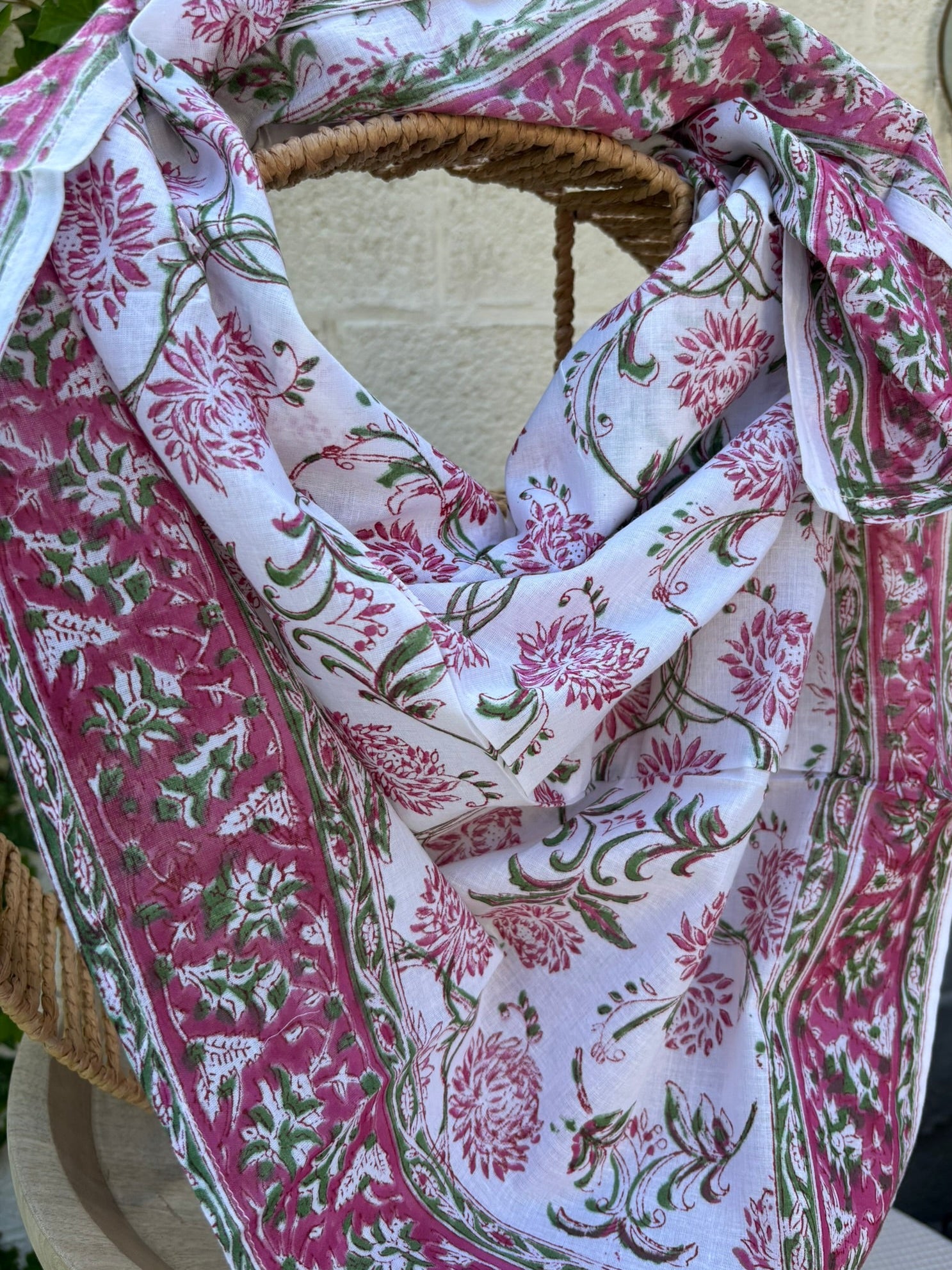 Un foulard en voile de coton blanc, léger et aérien, est présenté drapé sur une chaise en osier. Il est orné d'un imprimé artisanal blockprint représentant des fleurs de lotus et de chrysanthèmes stylisées dans des tons rose fuchsia et rose indien, reliées par des tiges et des feuilles vert tendre. Une large bordure fleurie assortie encadre la pièce. La mise en scène, avec un arrière-plan flou composé de lierre vert et d'un mur clair, souligne le caractère naturel, lumineux et bohème de cet accessoire