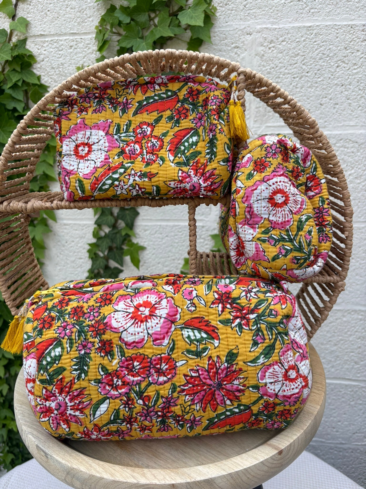 Un trio de trousses de maquillage ou pochettes assorties, en coton matelassé, est présenté sur une chaise en osier tissé. L'ensemble comprend une grande pochette plate, une trousse de toilette cylindrique et une plus petite pochette d'appoint. Toutes trois arborent un imprimé floral vibrant et dense dans des tons de rose fuchsia, rouge, blanc et vert sur un fond jaune moutarde lumineux. Deux des trousses sont ornées de glands jaunes décoratifs attachés aux tirettes de fermeture éclair. 
