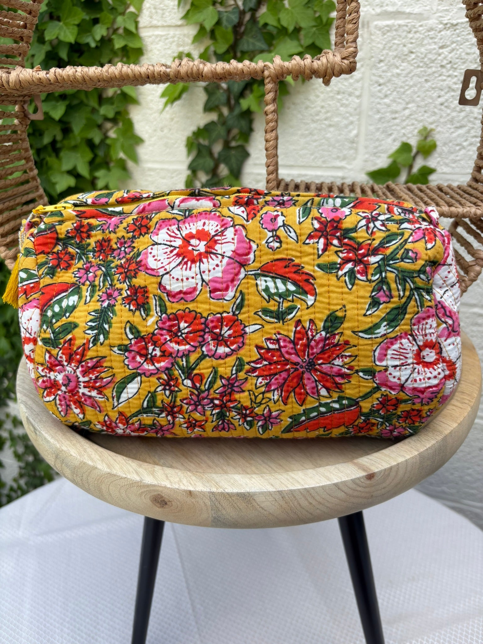 Trousse de maquillage ou pochette assortie, en coton matelassé, est présenté sur une chaise en osier tissé. Elle arbore un imprimé floral vibrant et dense dans des tons de rose fuchsia, rouge, blanc et vert sur un fond jaune moutarde lumineux. 