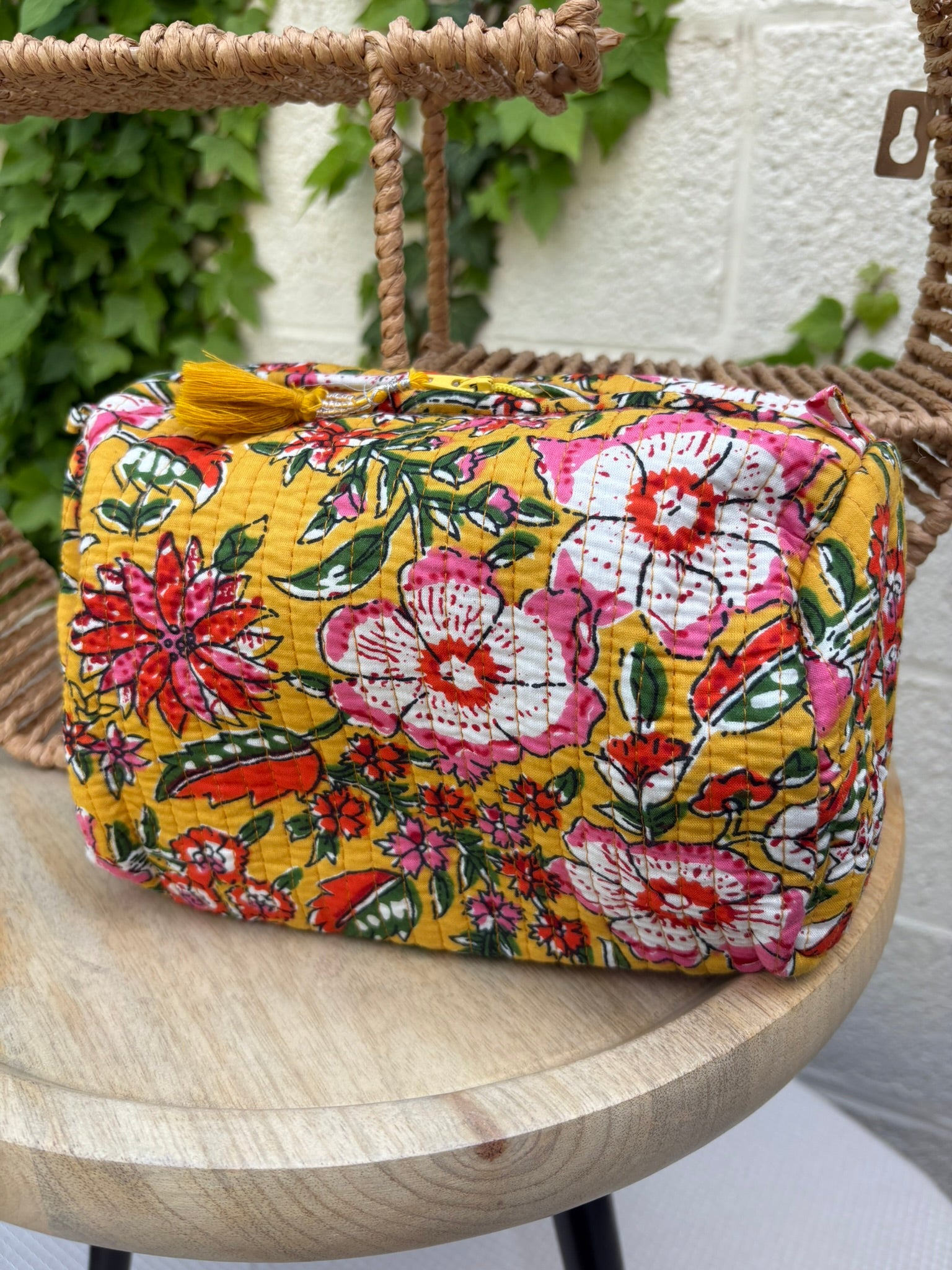 Trousse de maquillage ou pochette assortie, en coton matelassé, est présenté sur une chaise en osier tissé. Elle arbore un imprimé floral vibrant et dense dans des tons de rose fuchsia, rouge, blanc et vert sur un fond jaune moutarde lumineux. 