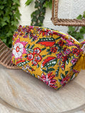 Trousse de maquillage ou pochette assortie, en coton matelassé, est présenté sur une chaise en osier tissé. Elle arbore un imprimé floral vibrant et dense dans des tons de rose fuchsia, rouge, blanc et vert sur un fond jaune moutarde lumineux. 