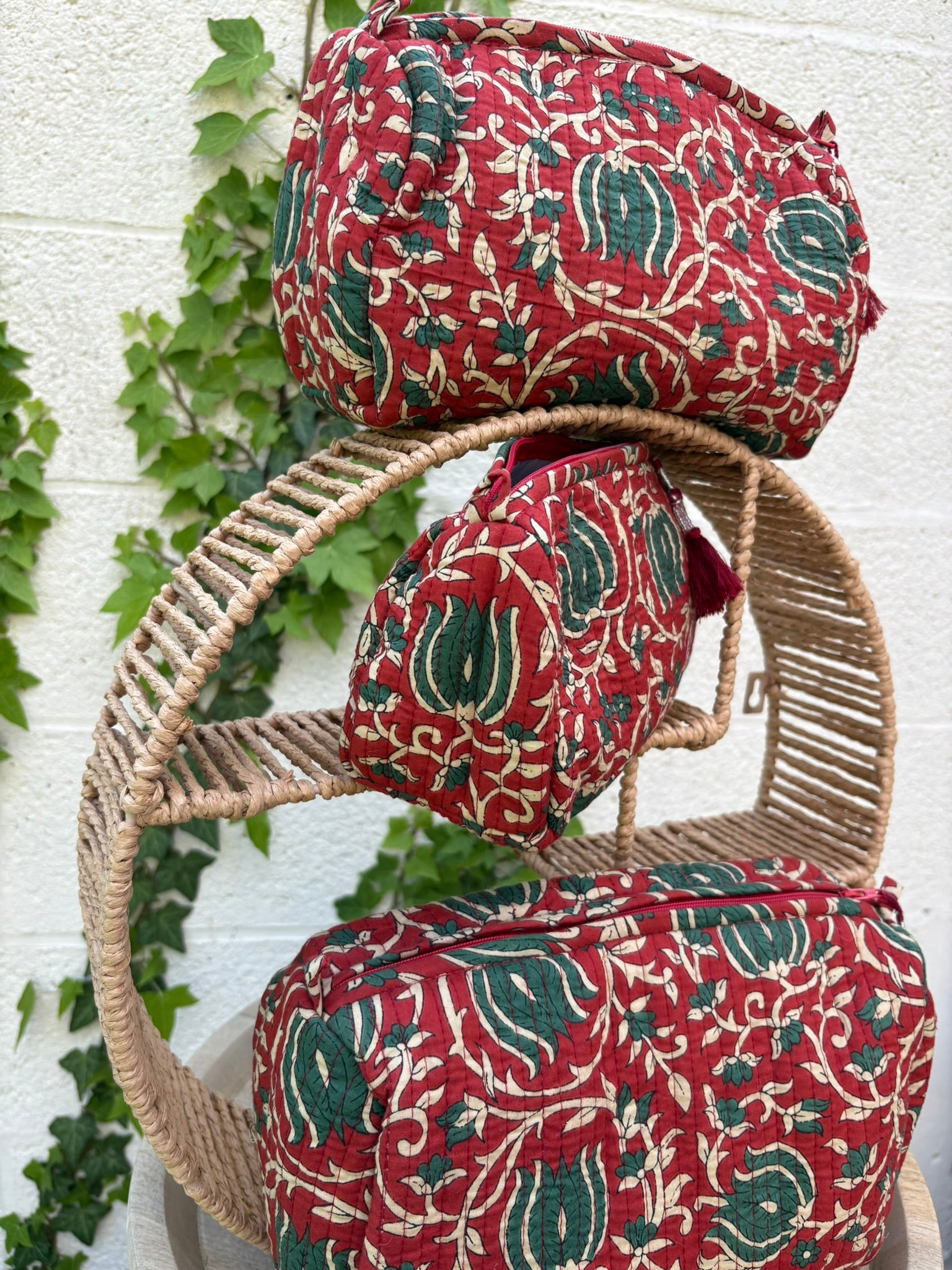 Trois trousses de toilette volumineuses en coton matelassé, ornées d'un imprimé blockprint traditionnel. Le motif floral complexe présente des teintes vert sapin et crème sur un fond rouge bordeaux profond. Les trousses sont parcourues de lignes de surpiqûres verticales (technique Kantha) qui ajoutent du relief et de la texture. Un pompon décoratif en fil rouge est attaché à la fermeture éclair. Elle est posée sur une surface blanche texturée, avec un mur clair et du lierre grimpant en arrière-plan