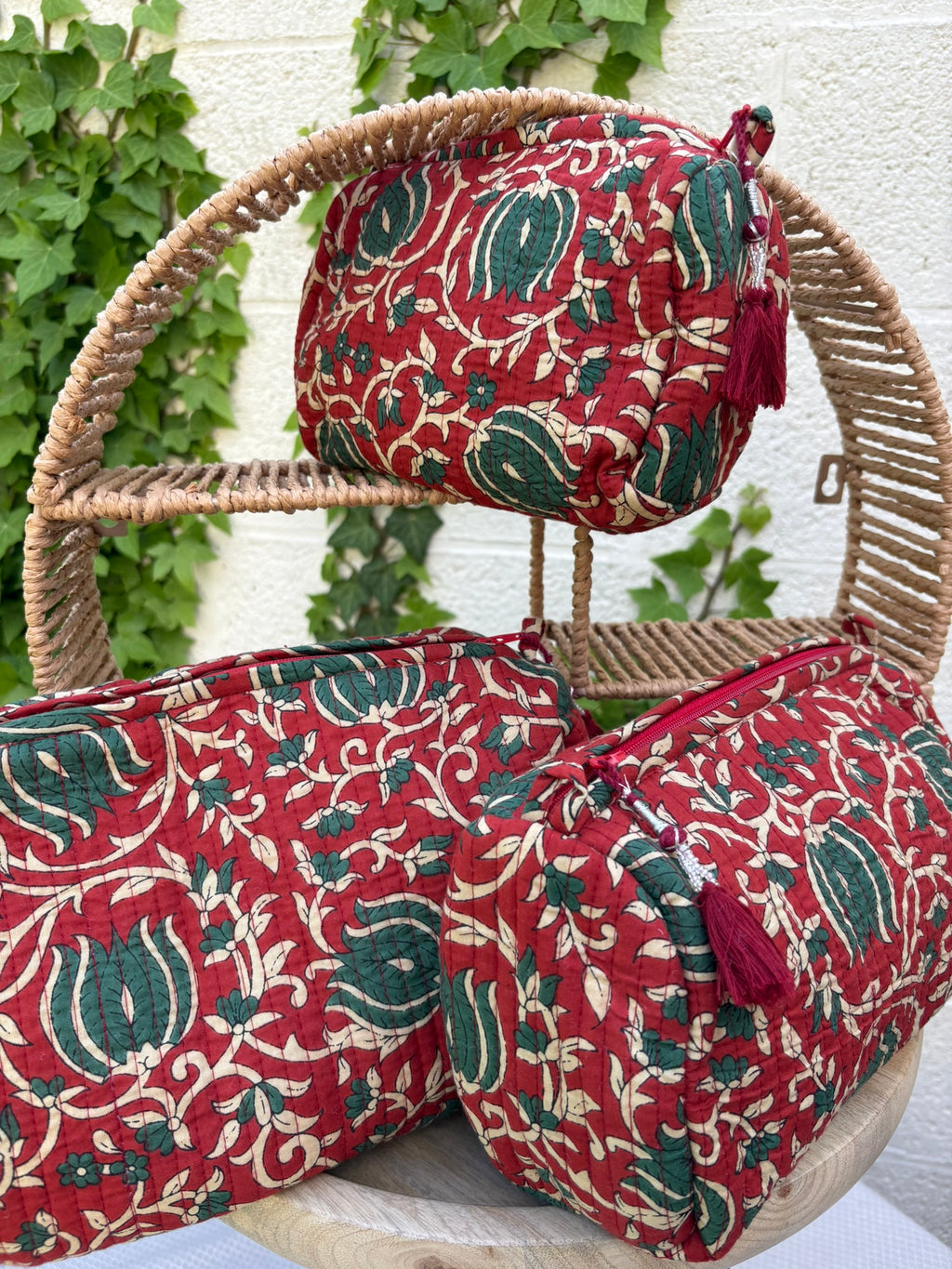 Trois trousses de toilette volumineuses en coton matelassé, ornées d'un imprimé blockprint traditionnel. Le motif floral complexe présente des teintes vert sapin et crème sur un fond rouge bordeaux profond. Les trousses sont parcourues de lignes de surpiqûres verticales (technique Kantha) qui ajoutent du relief et de la texture. Un pompon décoratif en fil rouge est attaché à la fermeture éclair. Elle est posée sur une surface blanche texturée, avec un mur clair et du lierre grimpant en arrière-plan