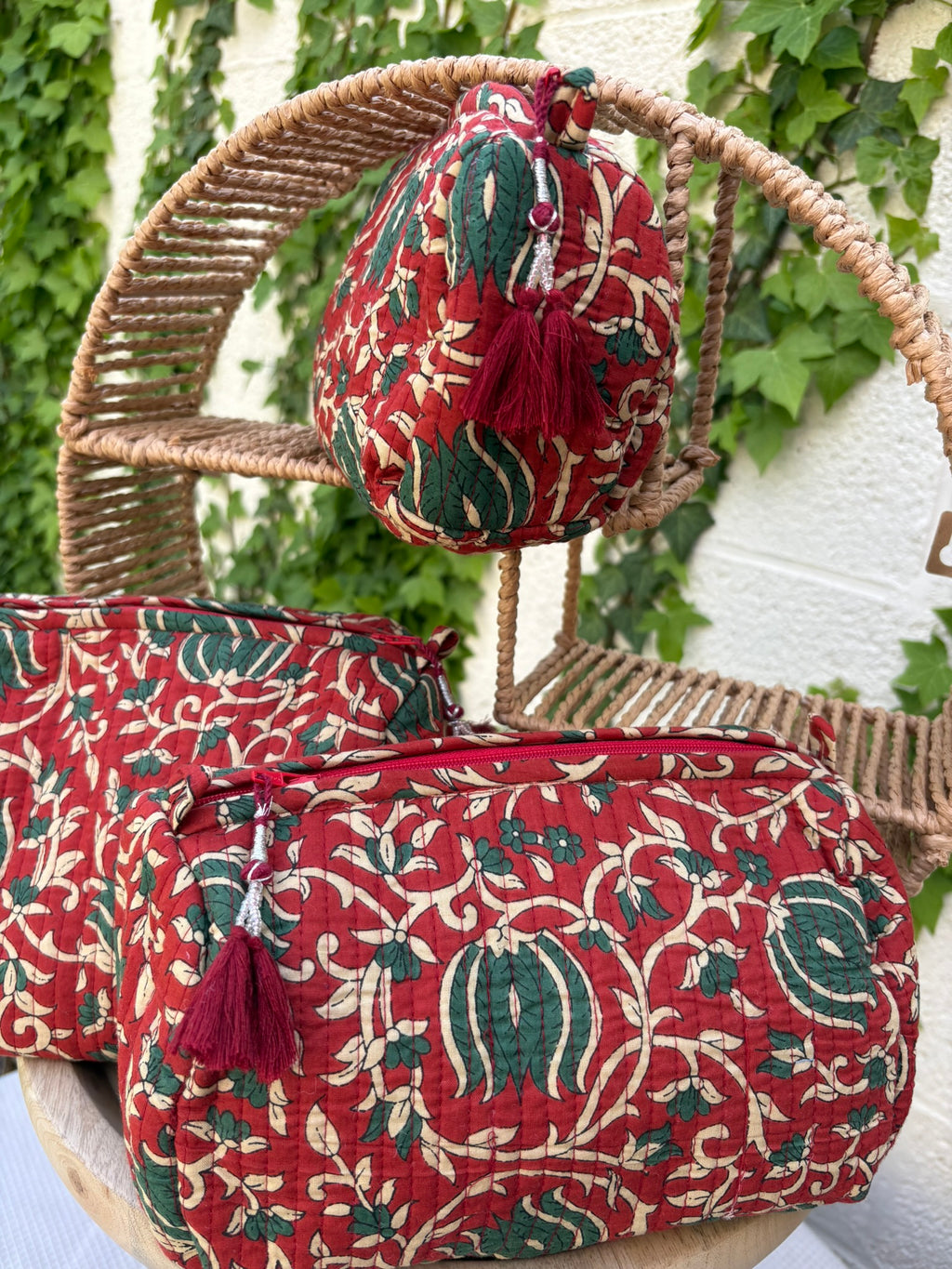 Trois trousses de toilette volumineuses en coton matelassé, ornées d'un imprimé blockprint traditionnel. Le motif floral complexe présente des teintes vert sapin et crème sur un fond rouge bordeaux profond. Les trousses sont parcourues de lignes de surpiqûres verticales (technique Kantha) qui ajoutent du relief et de la texture. Un pompon décoratif en fil rouge est attaché à la fermeture éclair. Elle est posée sur une surface blanche texturée, avec un mur clair et du lierre grimpant en arrière-plan