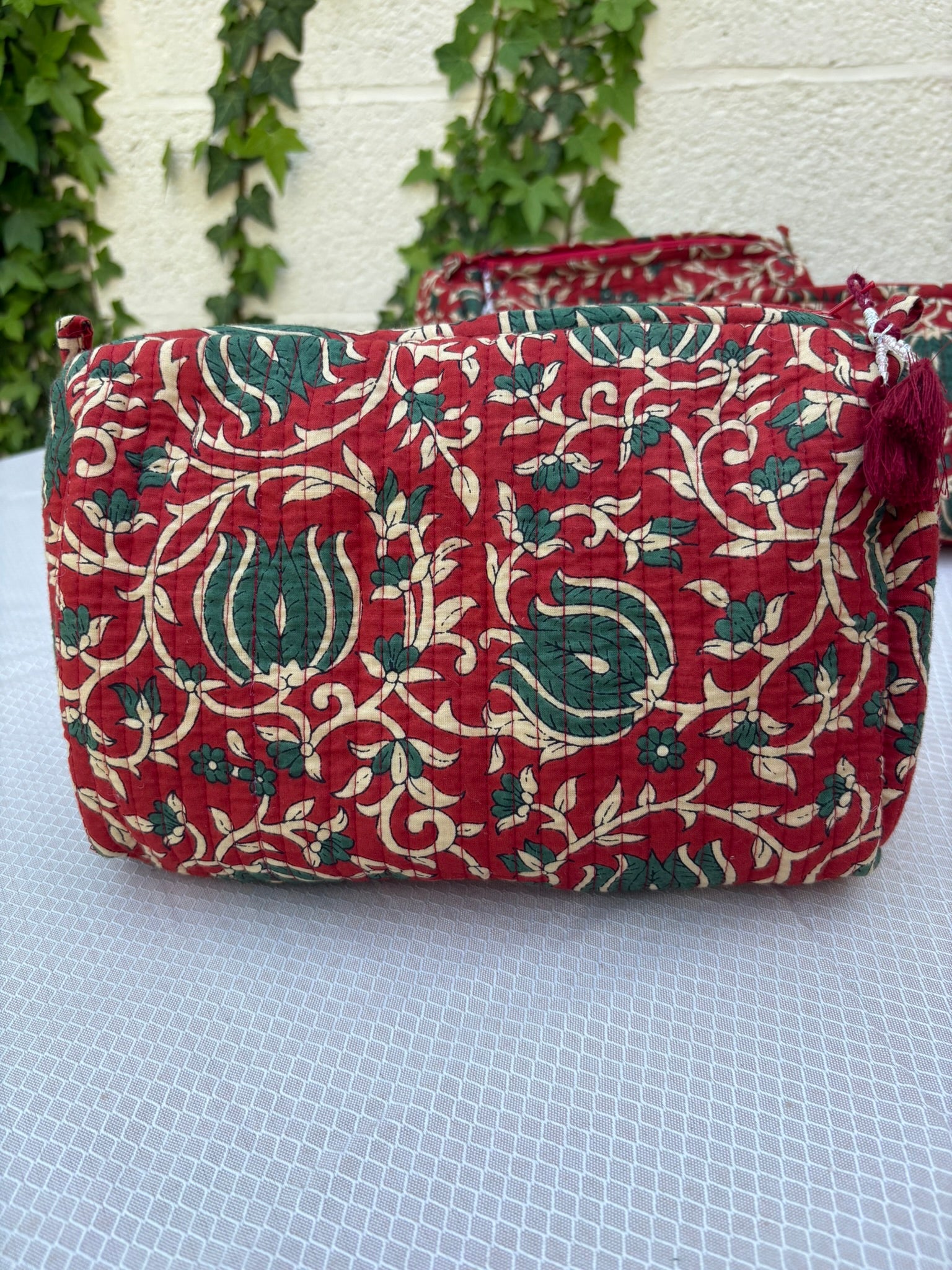Une trousse de toilette volumineuse en coton matelassé, ornée d'un imprimé blockprint traditionnel. Le motif floral complexe présente des teintes vert sapin et crème sur un fond rouge bordeaux profond. La trousse est parcourue de lignes de surpiqûres verticales (technique Kantha) qui ajoutent du relief et de la texture. Un pompon décoratif en fil rouge est attaché à la fermeture éclair. Elle est posée sur une surface blanche texturée, avec un mur clair et du lierre grimpant en arrière-plan