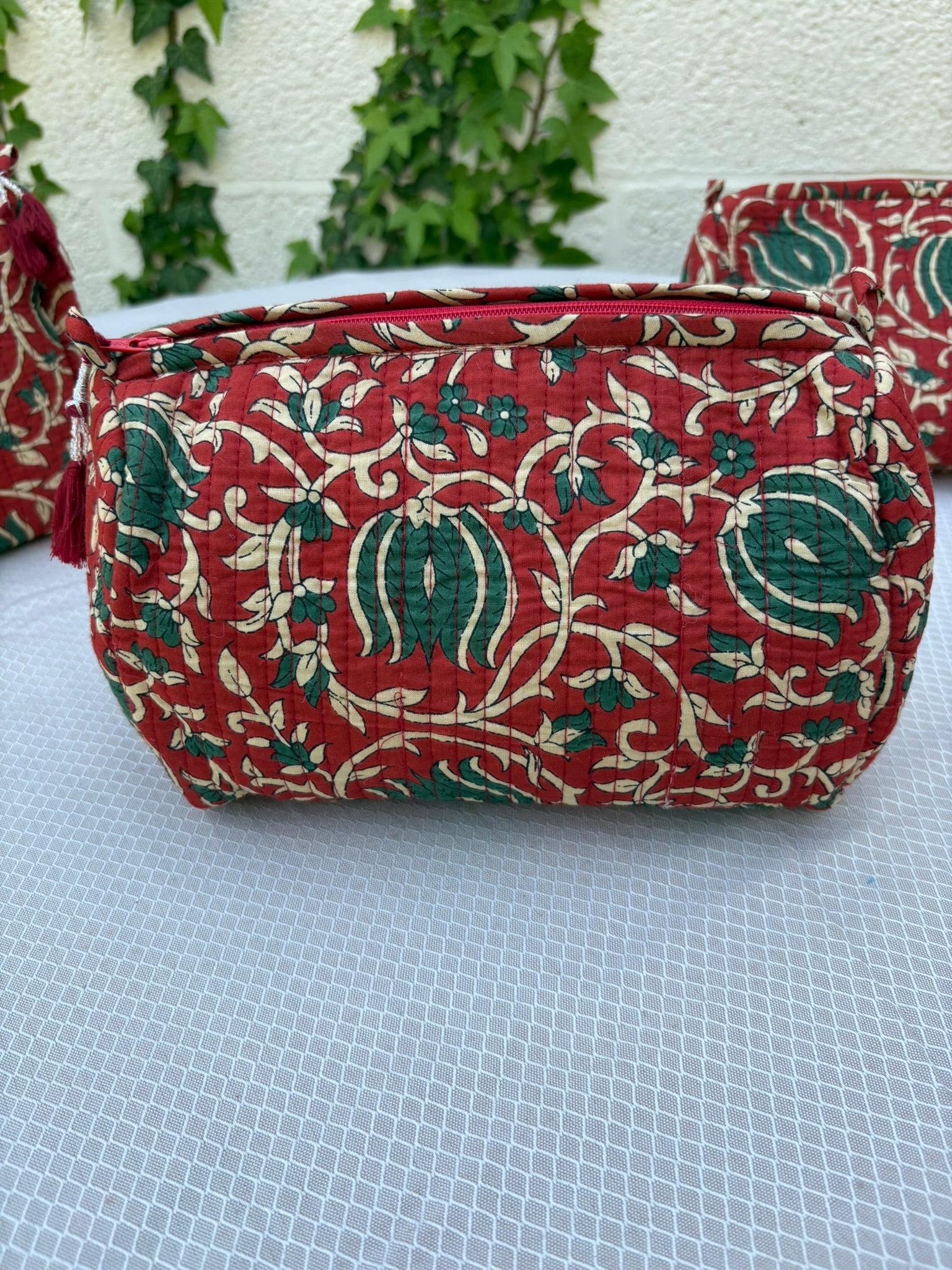 Une trousse de toilette volumineuse en coton matelassé, ornée d'un imprimé blockprint traditionnel. Le motif floral complexe présente des teintes vert sapin et crème sur un fond rouge bordeaux profond. La trousse est parcourue de lignes de surpiqûres verticales (technique Kantha) qui ajoutent du relief et de la texture. Un pompon décoratif en fil rouge est attaché à la fermeture éclair. Elle est posée sur une surface blanche texturée, avec un mur clair et du lierre grimpant en arrière-plan