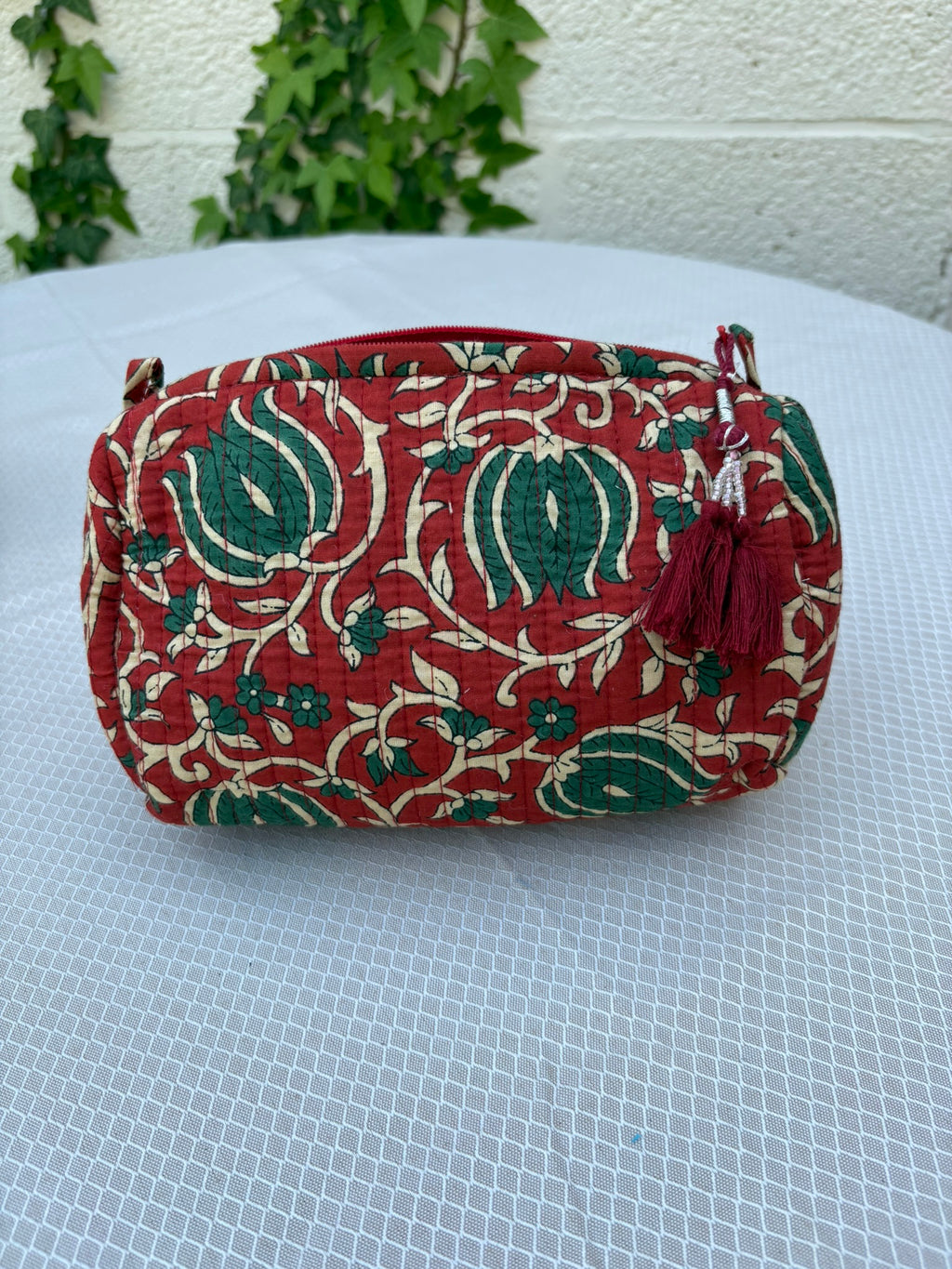 Une trousse de toilette volumineuse en coton matelassé, ornée d'un imprimé blockprint traditionnel. Le motif floral complexe présente des teintes vert sapin et crème sur un fond rouge bordeaux profond. La trousse est parcourue de lignes de surpiqûres verticales (technique Kantha) qui ajoutent du relief et de la texture. Un pompon décoratif en fil rouge est attaché à la fermeture éclair. Elle est posée sur une surface blanche texturée, avec un mur clair et du lierre grimpant en arrière-plan