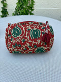 Une trousse de toilette volumineuse en coton matelassé, ornée d'un imprimé blockprint traditionnel. Le motif floral complexe présente des teintes vert sapin et crème sur un fond rouge bordeaux profond. La trousse est parcourue de lignes de surpiqûres verticales (technique Kantha) qui ajoutent du relief et de la texture. Un pompon décoratif en fil rouge est attaché à la fermeture éclair. Elle est posée sur une surface blanche texturée, avec un mur clair et du lierre grimpant en arrière-plan