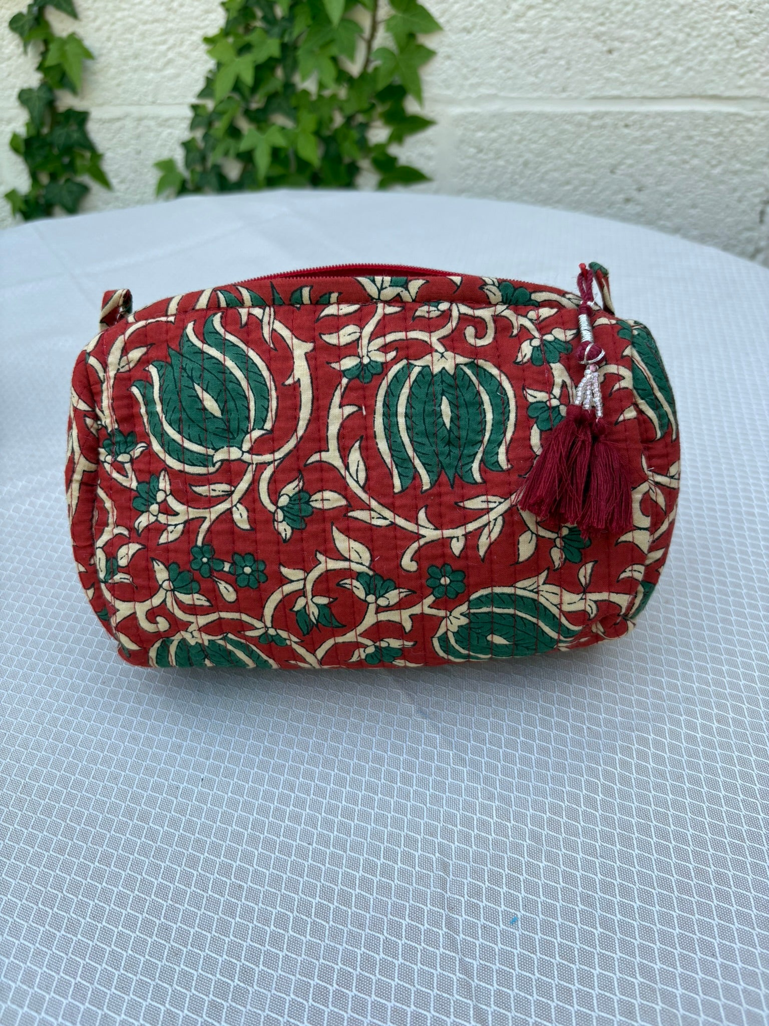 Une trousse de toilette volumineuse en coton matelassé, ornée d'un imprimé blockprint traditionnel. Le motif floral complexe présente des teintes vert sapin et crème sur un fond rouge bordeaux profond. La trousse est parcourue de lignes de surpiqûres verticales (technique Kantha) qui ajoutent du relief et de la texture. Un pompon décoratif en fil rouge est attaché à la fermeture éclair. Elle est posée sur une surface blanche texturée, avec un mur clair et du lierre grimpant en arrière-plan