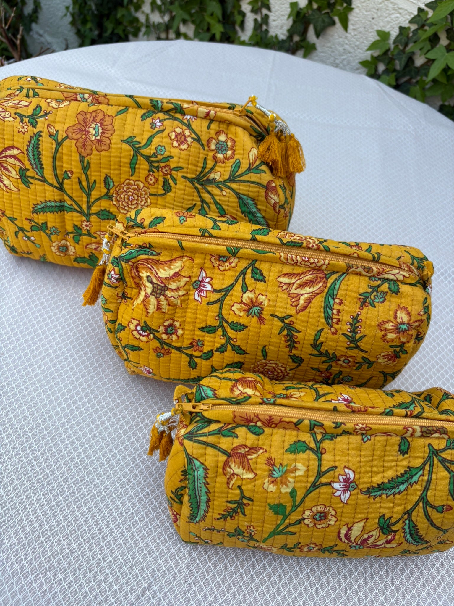 Trois trousses de toilette assorties en coton matelassé jaune safran sont présentées sur une étagère circulaire en osier tressé. Les trousses arborent un imprimé blockprint délicat composé de fleurs et de feuillages dans des tons de vert émeraude, turquoise, rouge bordeaux et blanc. La texture matelassée est soulignée par des surpiqûres verticales régulières (technique Kantha). Chaque trousse est équipée d'une fermeture éclair jaune ornée d'un pompon décoratif assorti. 
