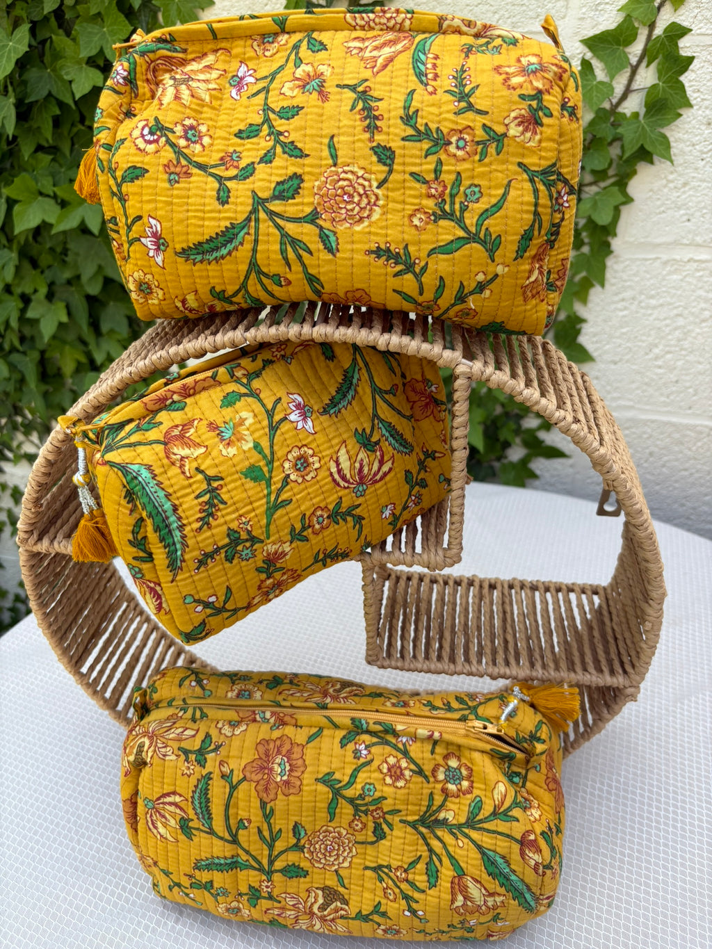 Trois trousses de toilette assorties en coton matelassé jaune safran sont présentées sur une étagère circulaire en osier tressé. Les trousses arborent un imprimé blockprint délicat composé de fleurs et de feuillages dans des tons de vert émeraude, turquoise, rouge bordeaux et blanc. La texture matelassée est soulignée par des surpiqûres verticales régulières (technique Kantha). Chaque trousse est équipée d'une fermeture éclair jaune ornée d'un pompon décoratif assorti. 