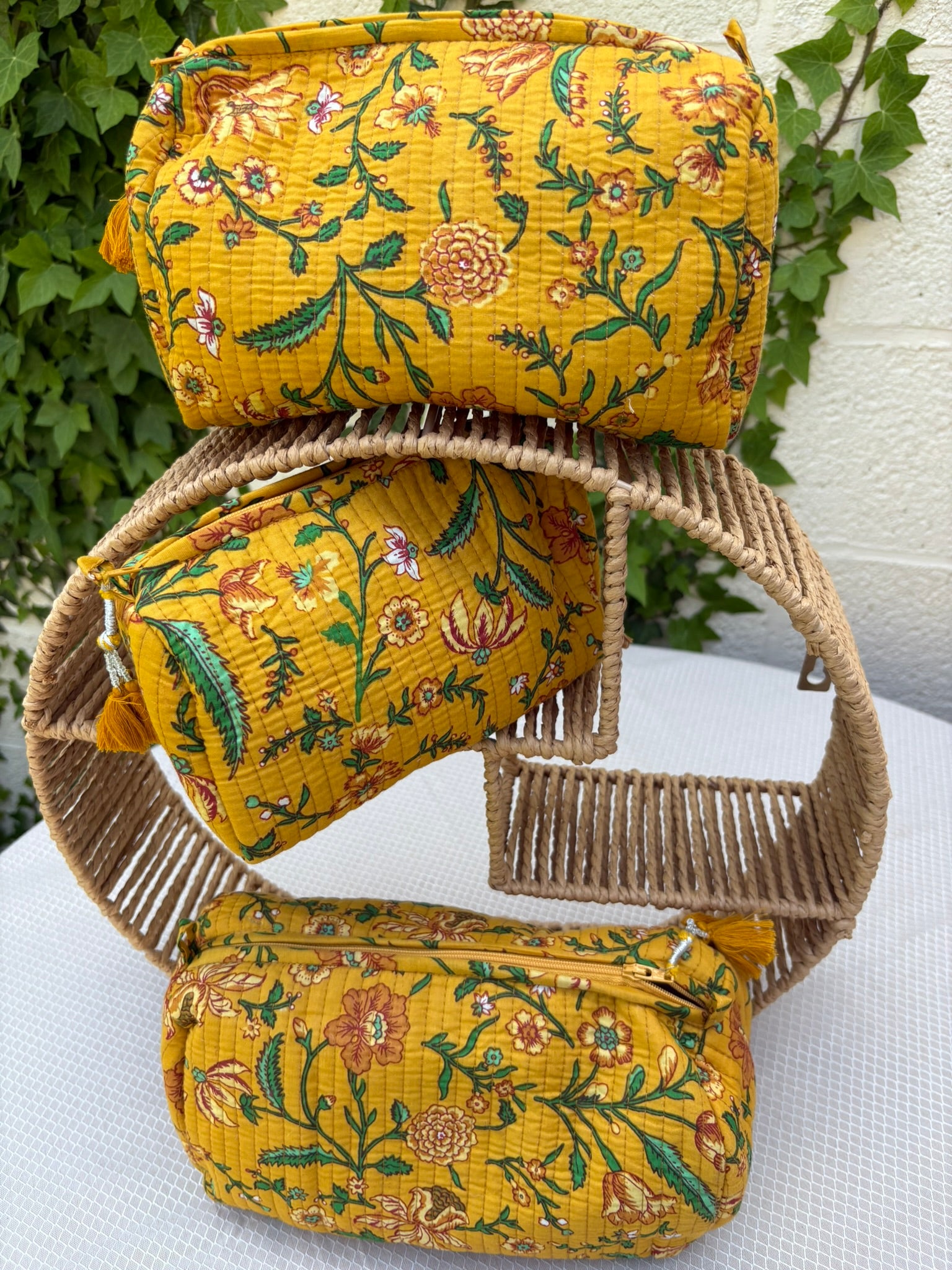 Trois trousses de toilette assorties en coton matelassé jaune safran sont présentées sur une étagère circulaire en osier tressé. Les trousses arborent un imprimé blockprint délicat composé de fleurs et de feuillages dans des tons de vert émeraude, turquoise, rouge bordeaux et blanc. La texture matelassée est soulignée par des surpiqûres verticales régulières (technique Kantha). Chaque trousse est équipée d'une fermeture éclair jaune ornée d'un pompon décoratif assorti. 