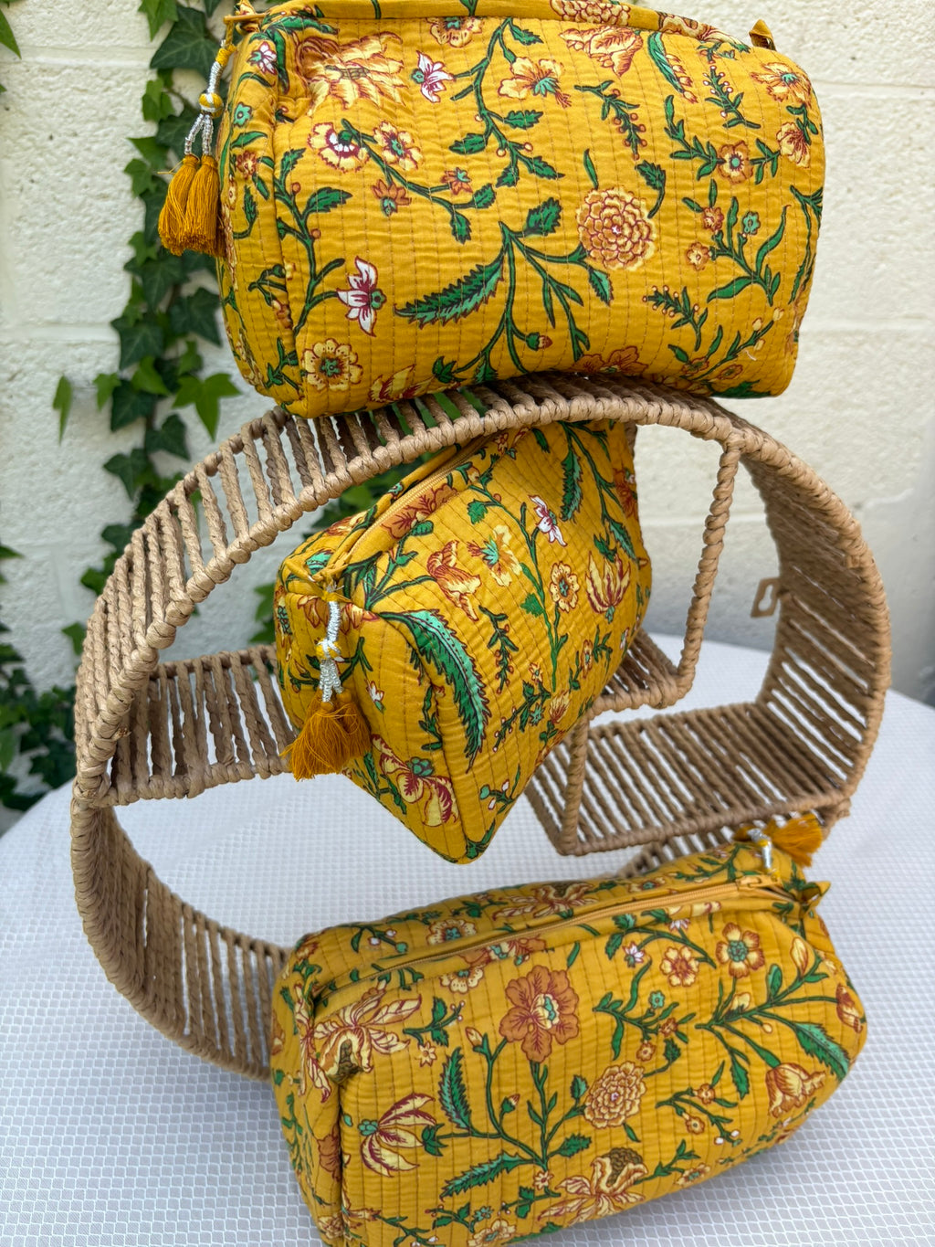 Trois trousses de toilette assorties en coton matelassé jaune safran sont présentées sur une étagère circulaire en osier tressé. Les trousses arborent un imprimé blockprint délicat composé de fleurs et de feuillages dans des tons de vert émeraude, turquoise, rouge bordeaux et blanc. La texture matelassée est soulignée par des surpiqûres verticales régulières (technique Kantha). Chaque trousse est équipée d'une fermeture éclair jaune ornée d'un pompon décoratif assorti. 