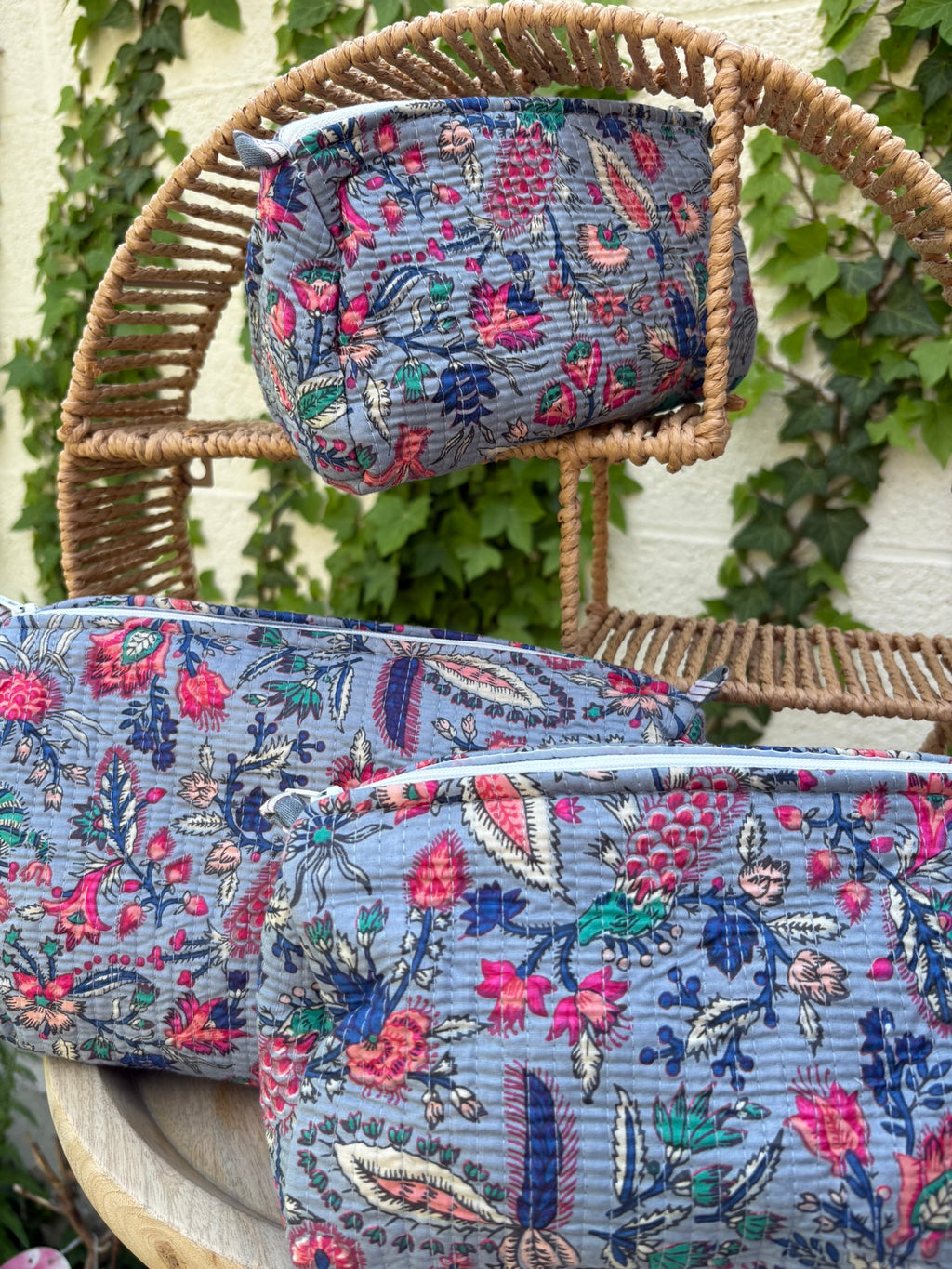 Un ensemble de trois trousses de toilette de tailles différentes, confectionnées en coton matelassé selon la technique traditionnelle Kantha, est exposé de façon artistique sur une étagère circulaire en osier. Le tissu présente un imprimé blockprint dense avec des motifs floraux et végétaux multicolores (rose fuchsia, turquoise,  et crème) sur un fond bleu grisâtre élégant. La texture est enrichie par des surpiqûres horizontales régulières. La scène est photographiée en extérieur avec un mur blanc 
