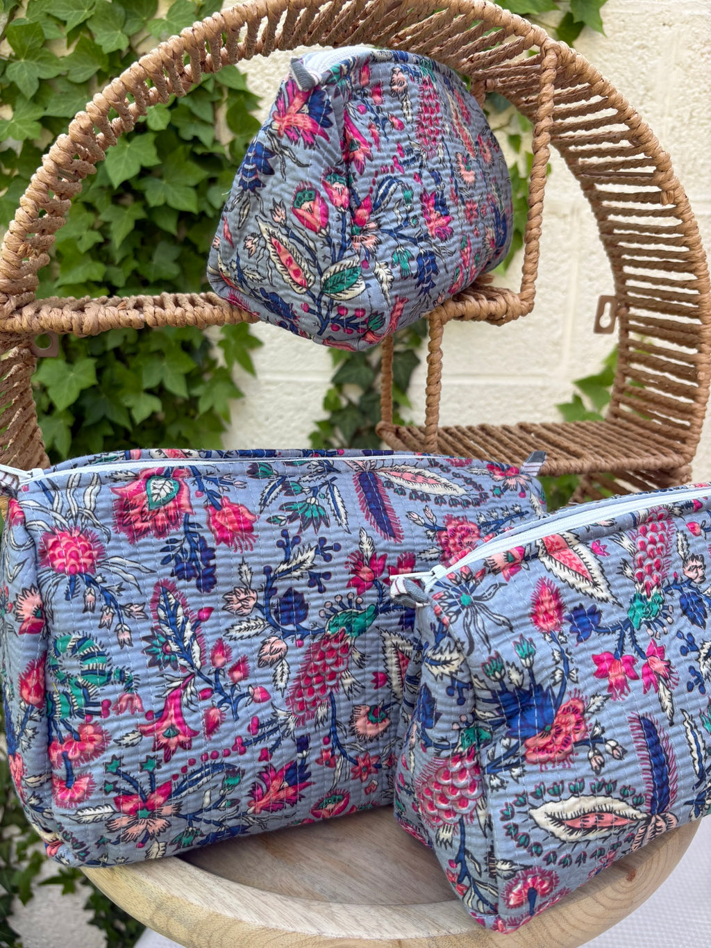 Un ensemble de trois trousses de toilette de tailles différentes, confectionnées en coton matelassé selon la technique traditionnelle Kantha, est exposé de façon artistique sur une étagère circulaire en osier. Le tissu présente un imprimé blockprint dense avec des motifs floraux et végétaux multicolores (rose fuchsia, turquoise,  et crème) sur un fond bleu grisâtre élégant. La texture est enrichie par des surpiqûres horizontales régulières. La scène est photographiée en extérieur avec un mur blanc 
