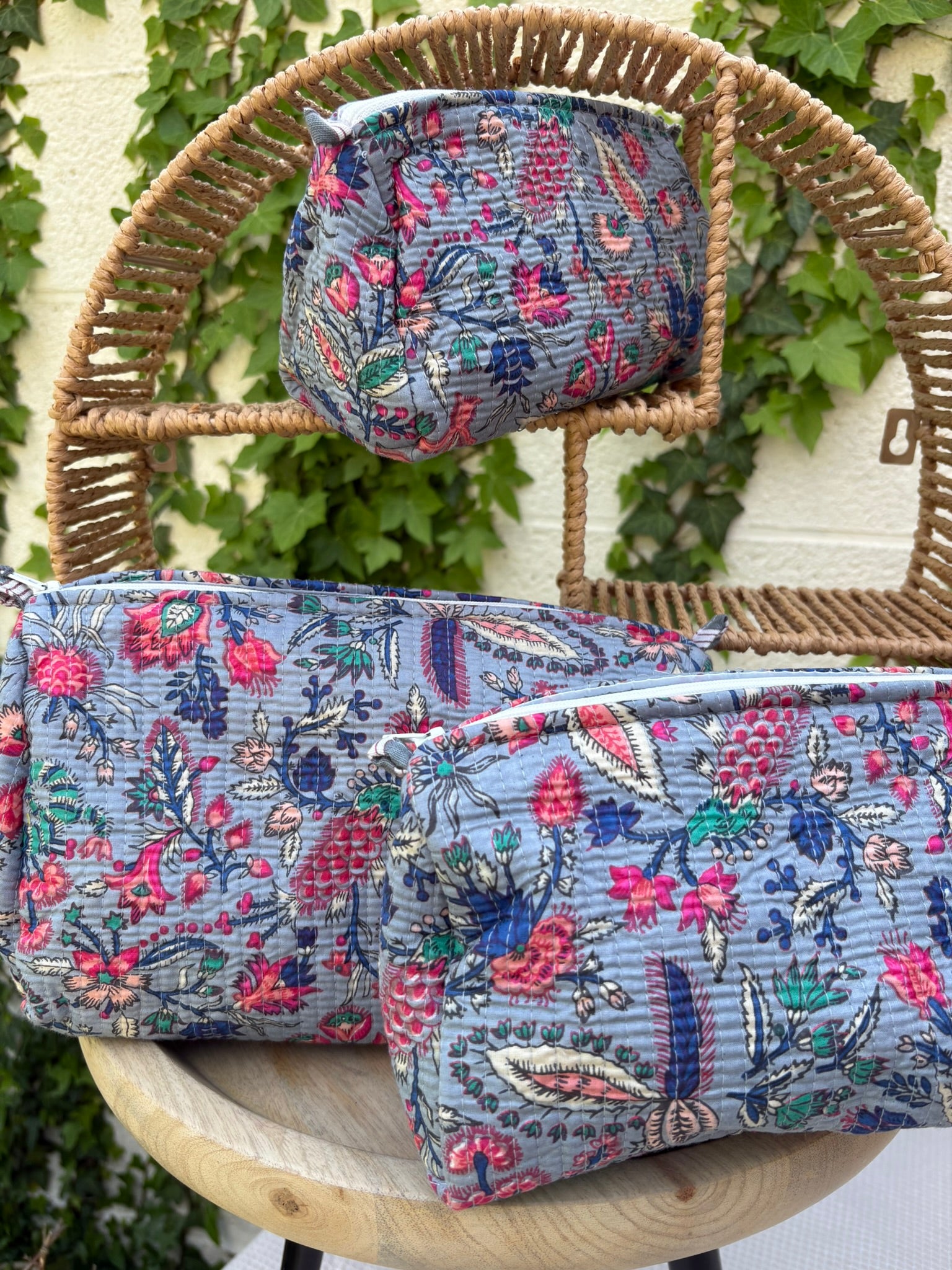 Un ensemble de trois trousses de toilette de tailles différentes, confectionnées en coton matelassé selon la technique traditionnelle Kantha, est exposé de façon artistique sur une étagère circulaire en osier. Le tissu présente un imprimé blockprint dense avec des motifs floraux et végétaux multicolores (rose fuchsia, turquoise,  et crème) sur un fond bleu grisâtre élégant. La texture est enrichie par des surpiqûres horizontales régulières. La scène est photographiée en extérieur avec un mur blanc 