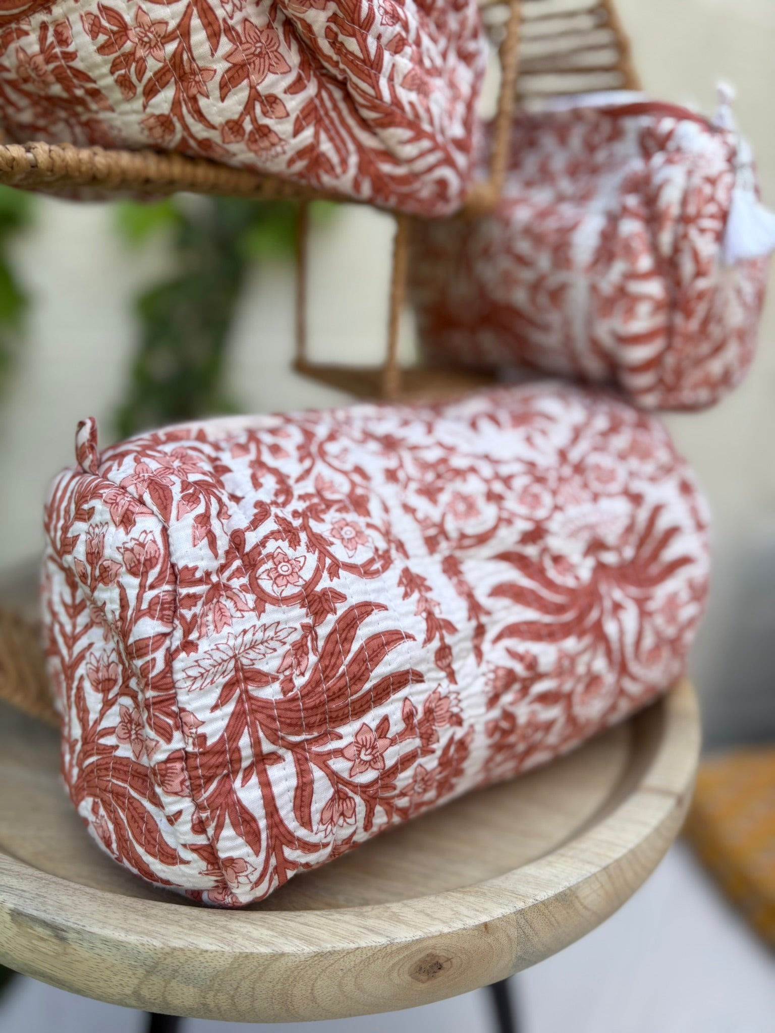 Un ensemble de trois trousses de toilette cylindriques en coton matelassé (technique Kantha) est présenté sur une étagère circulaire en osier tressé. Le tissu blanc est orné d'un imprimé blockprint monochrome dans des tons terre cuite et brique, représentant des motifs floraux et des feuillages denses. La texture est marquée par des surpiqûres horizontales régulières. La scène, capturée en extérieur avec un arrière-plan de lierre vert sur un mur clair, met en valeur le caractère artisanal et naturel 